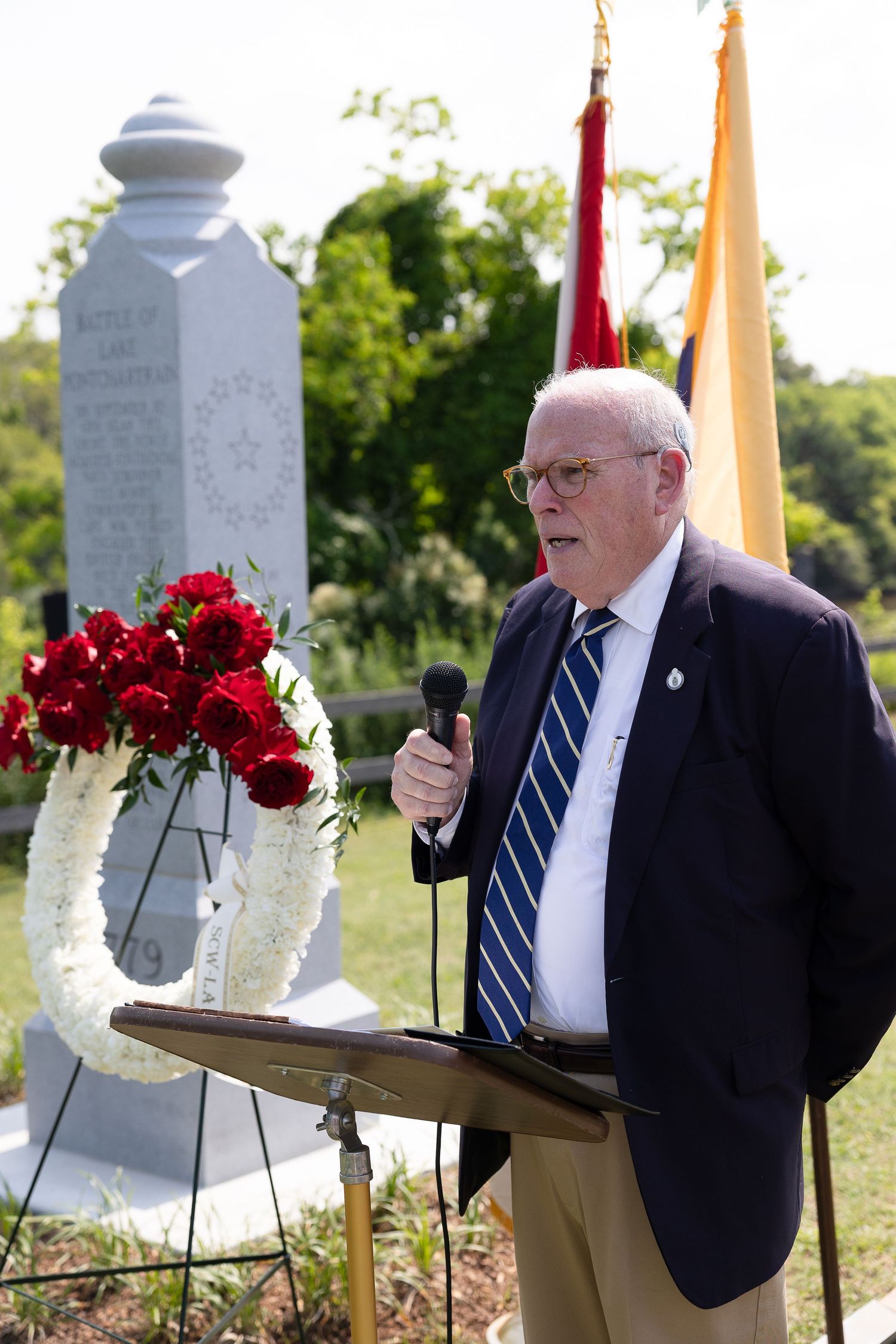 A speaker in a navy suit gestures while delivering remarks at a memorial dedication in Mandeville, Louisiana.