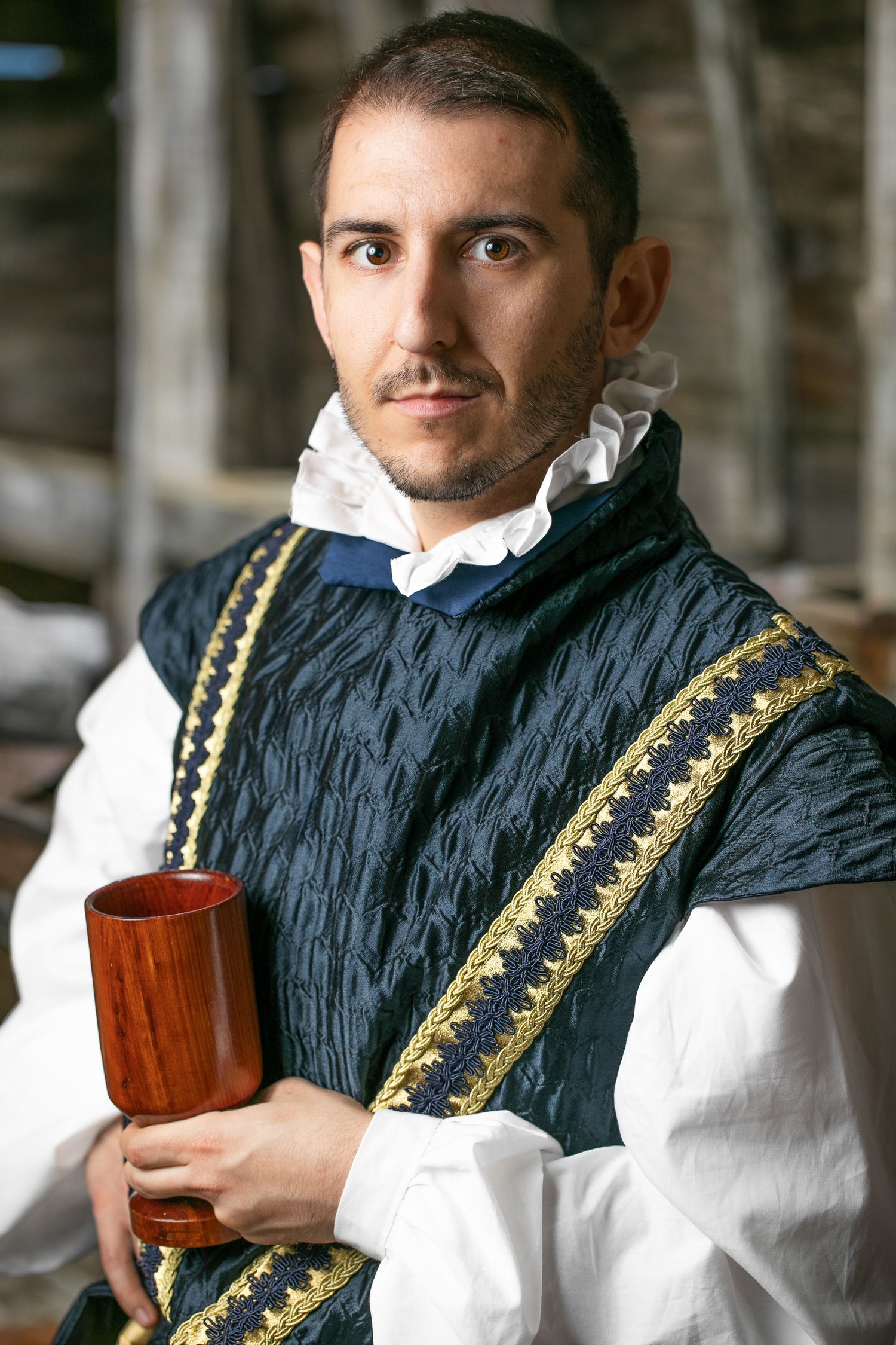 Renaissance style portrait showing a person in navy blue doublet with gold trim and white ruffled collar holding a wooden cup.