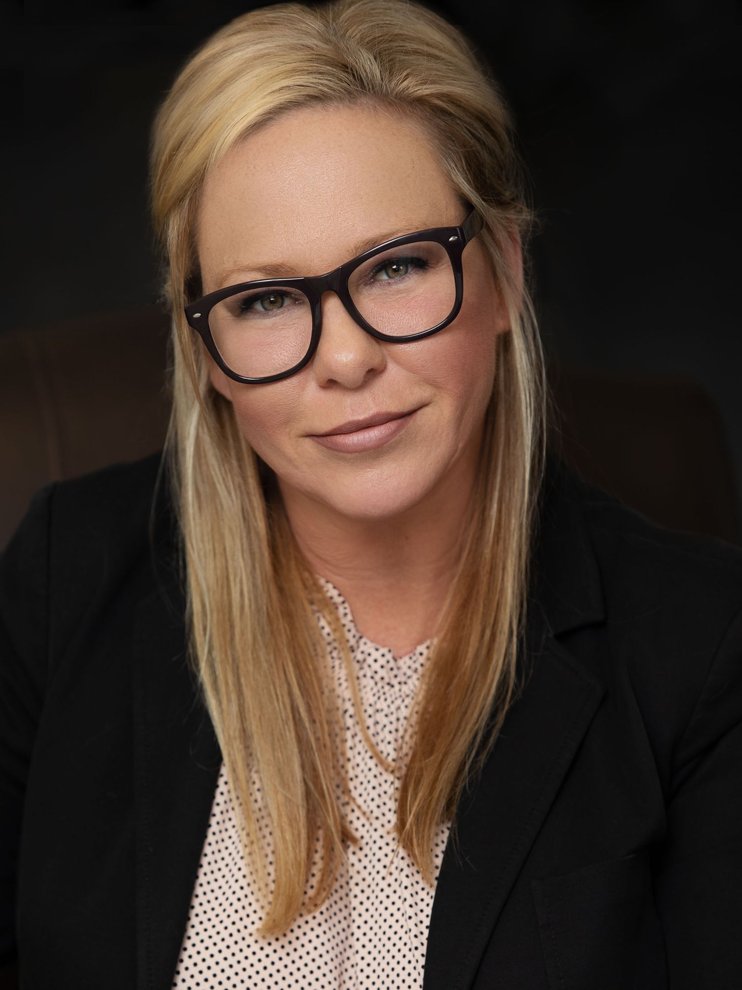 Professional business portrait of person wearing glasses and black blazer with blonde hair and polite smile against dark background.