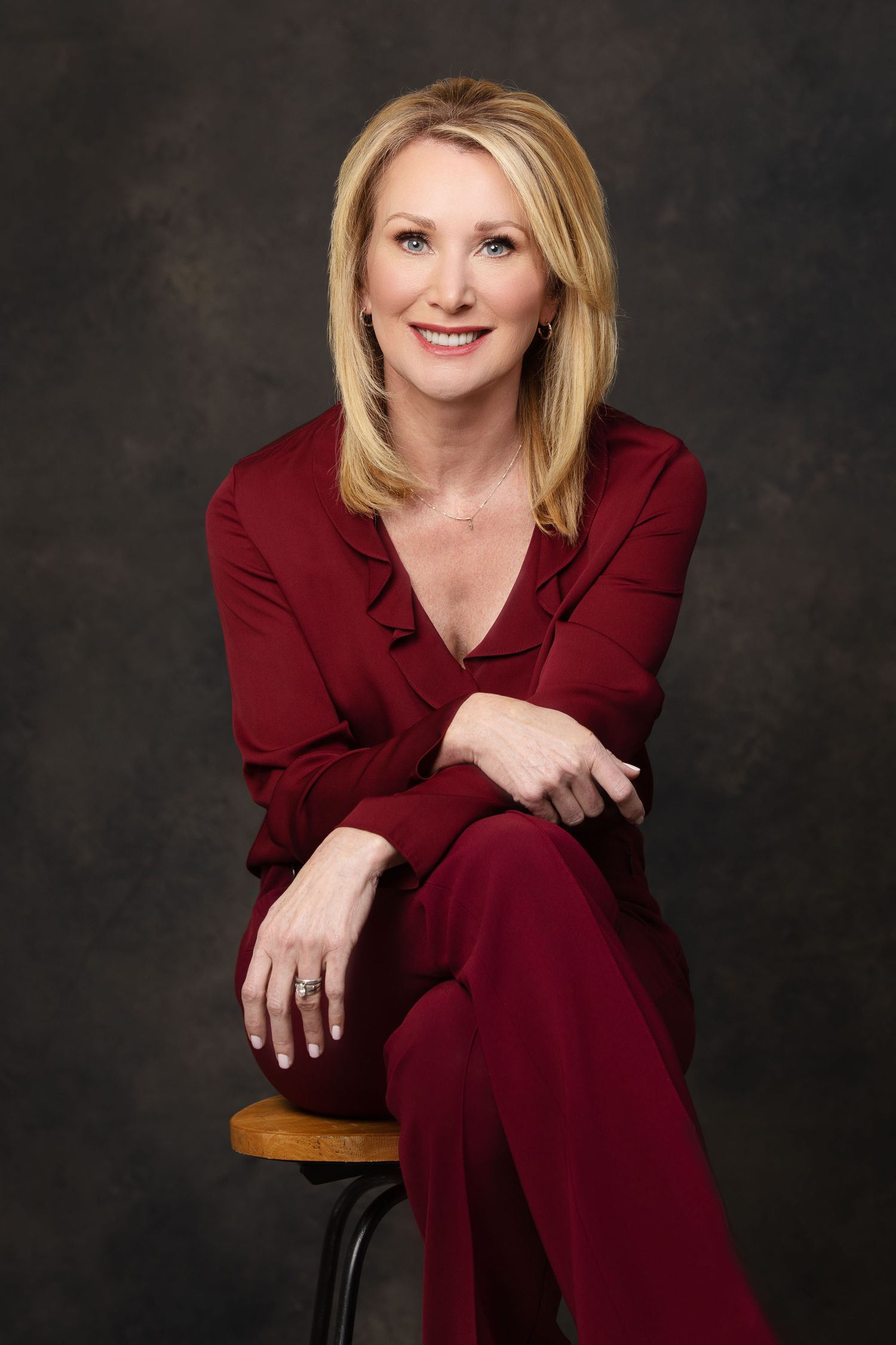 Professional studio portrait of a person seated on a stool wearing a burgundy pantsuit against a dark background.