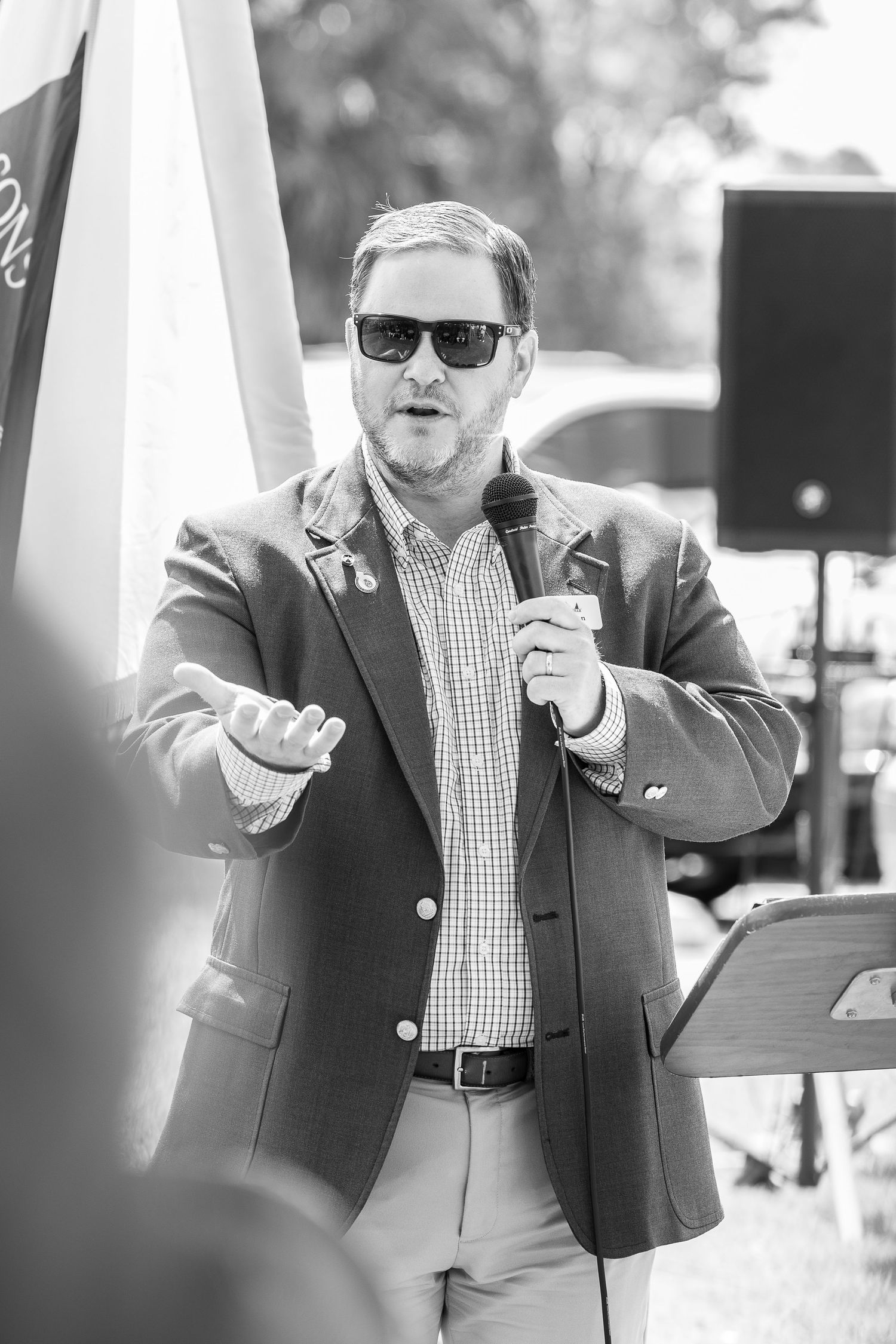 A speaker in a suit jacket and sunglasses gestures while holding a microphone during an outdoor event. Bellus event photography new orleans