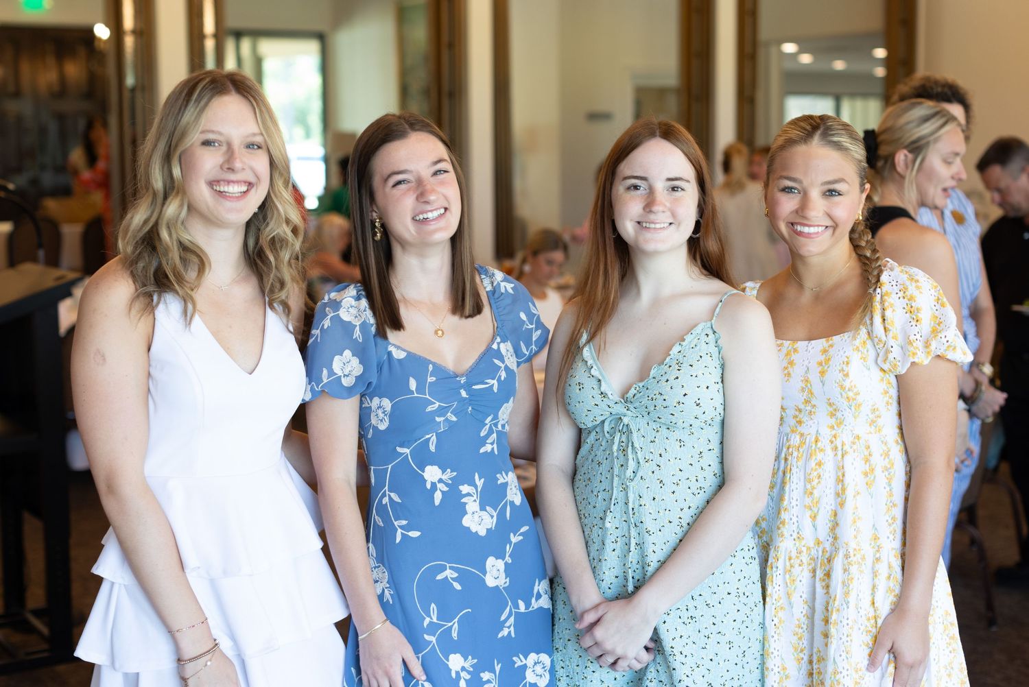 A group of women stand together smiling while wearing coordinated blue and white summer dresses at a special occasion.
