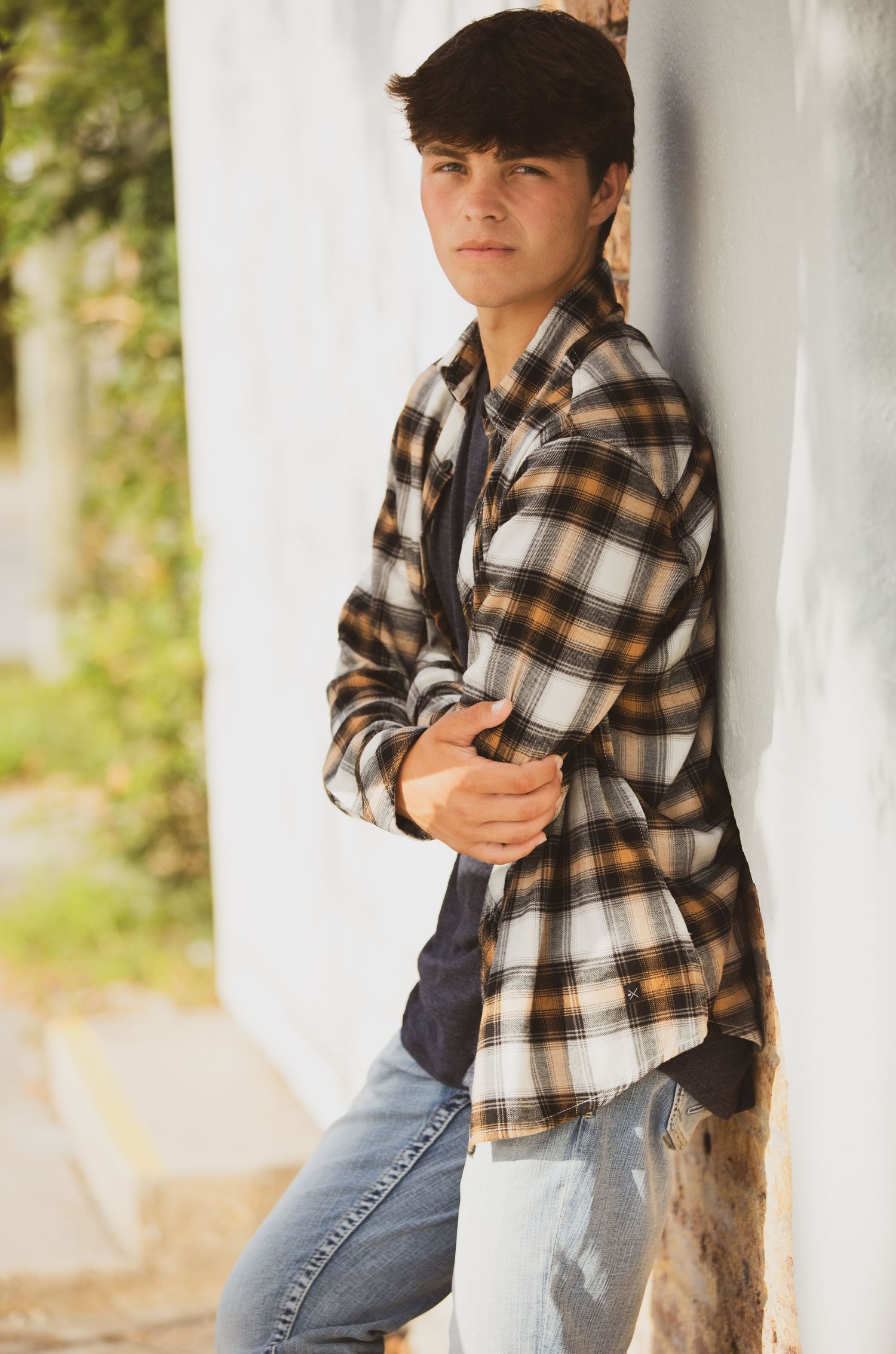 Child in brown plaid flannel shirt and jeans strikes casual poses while leaning against white wall outdoors.