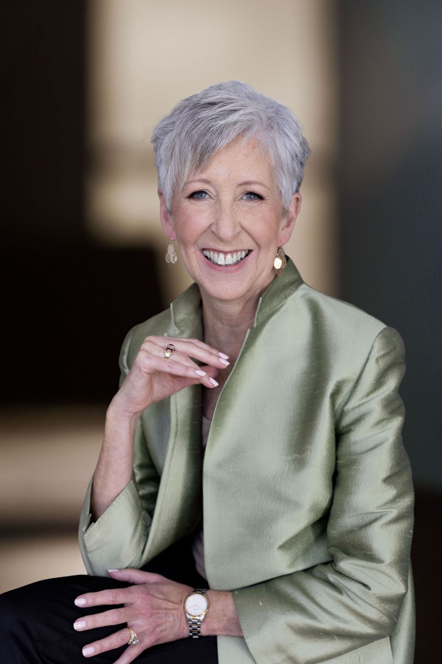 Professional portrait of someone wearing a sage green silk blazer and silver jewelry, with short gray hair and a warm smile.