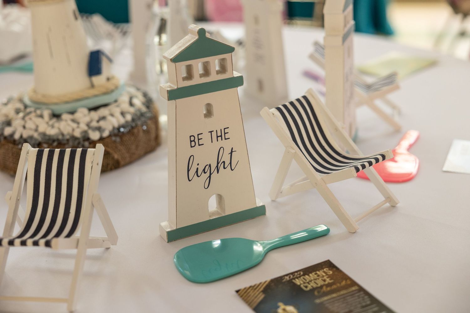Coastal themed table decor featuring a lighthouse sign reading 'Be the Light' with striped beach chairs and mint green accents.