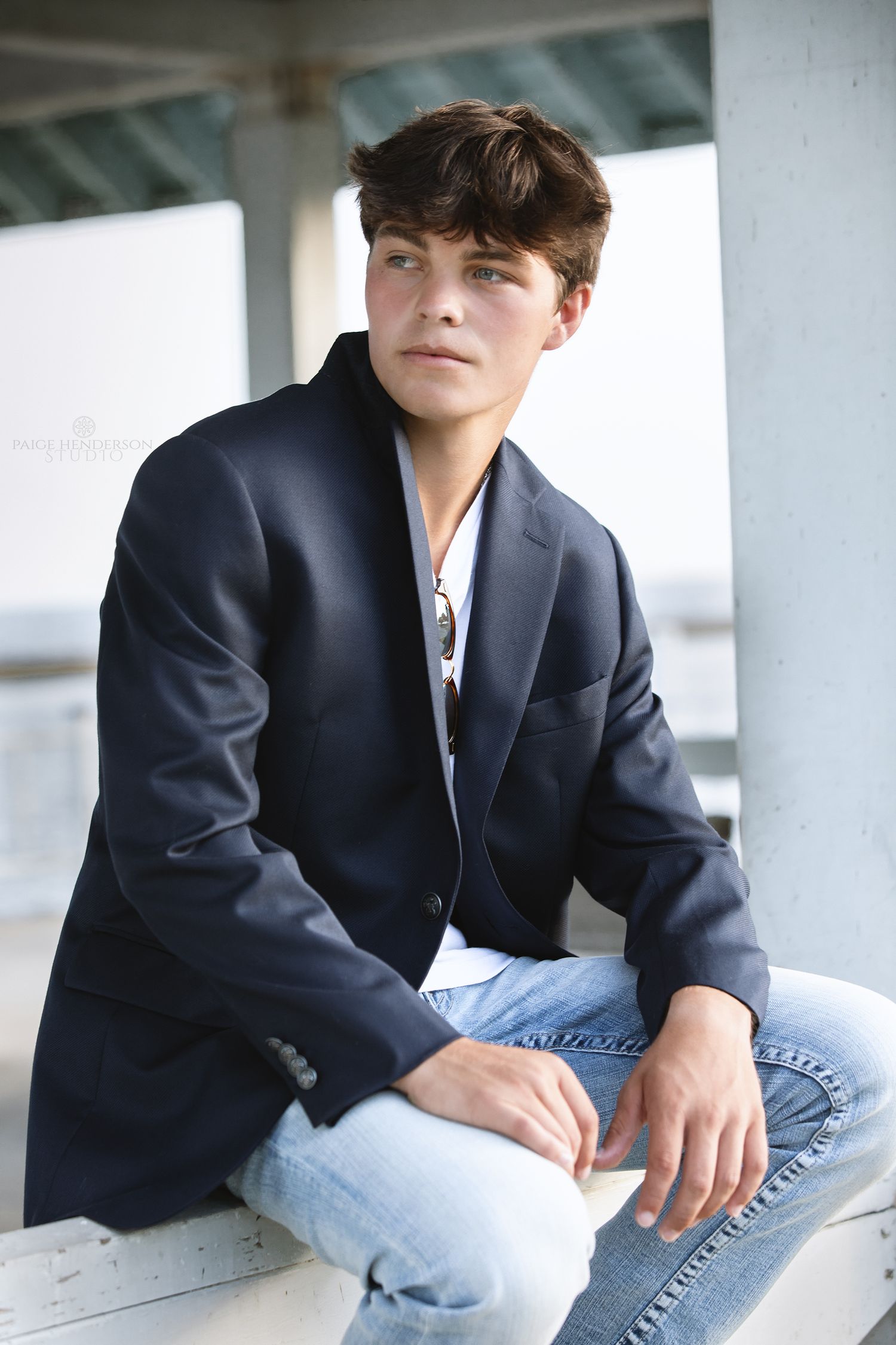 A young person in a navy blazer and jeans poses casually against a white surface in a portrait-style photo.