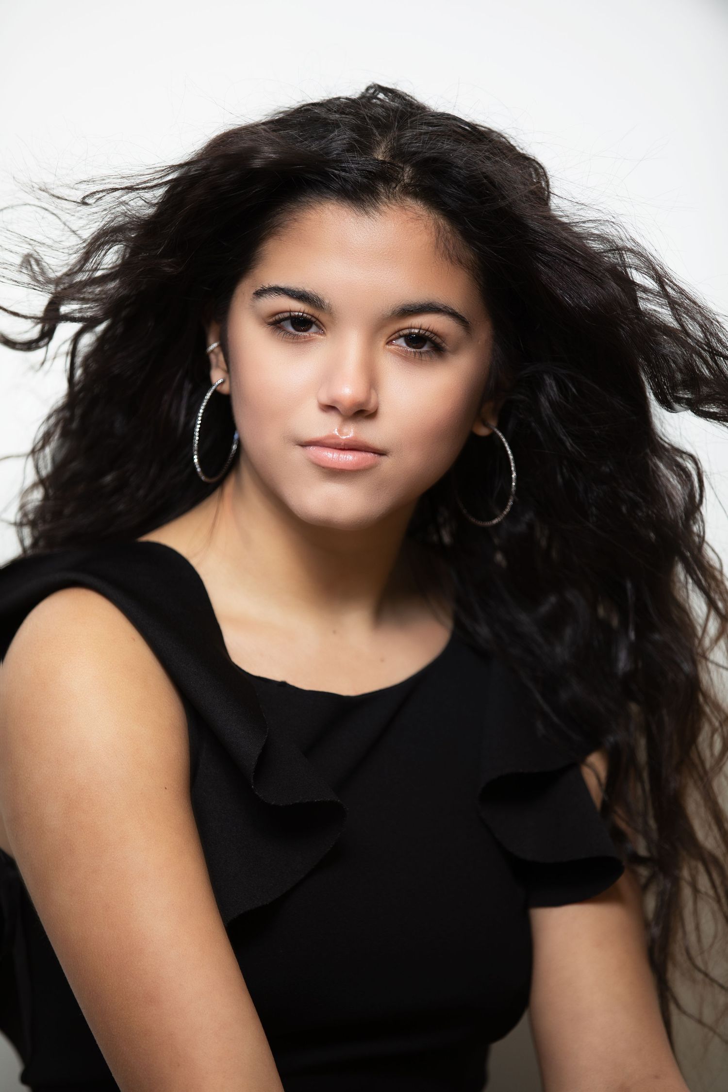 Portrait of a young woman in a black top with flowing dark hair and silver hoop earrings against a light background.