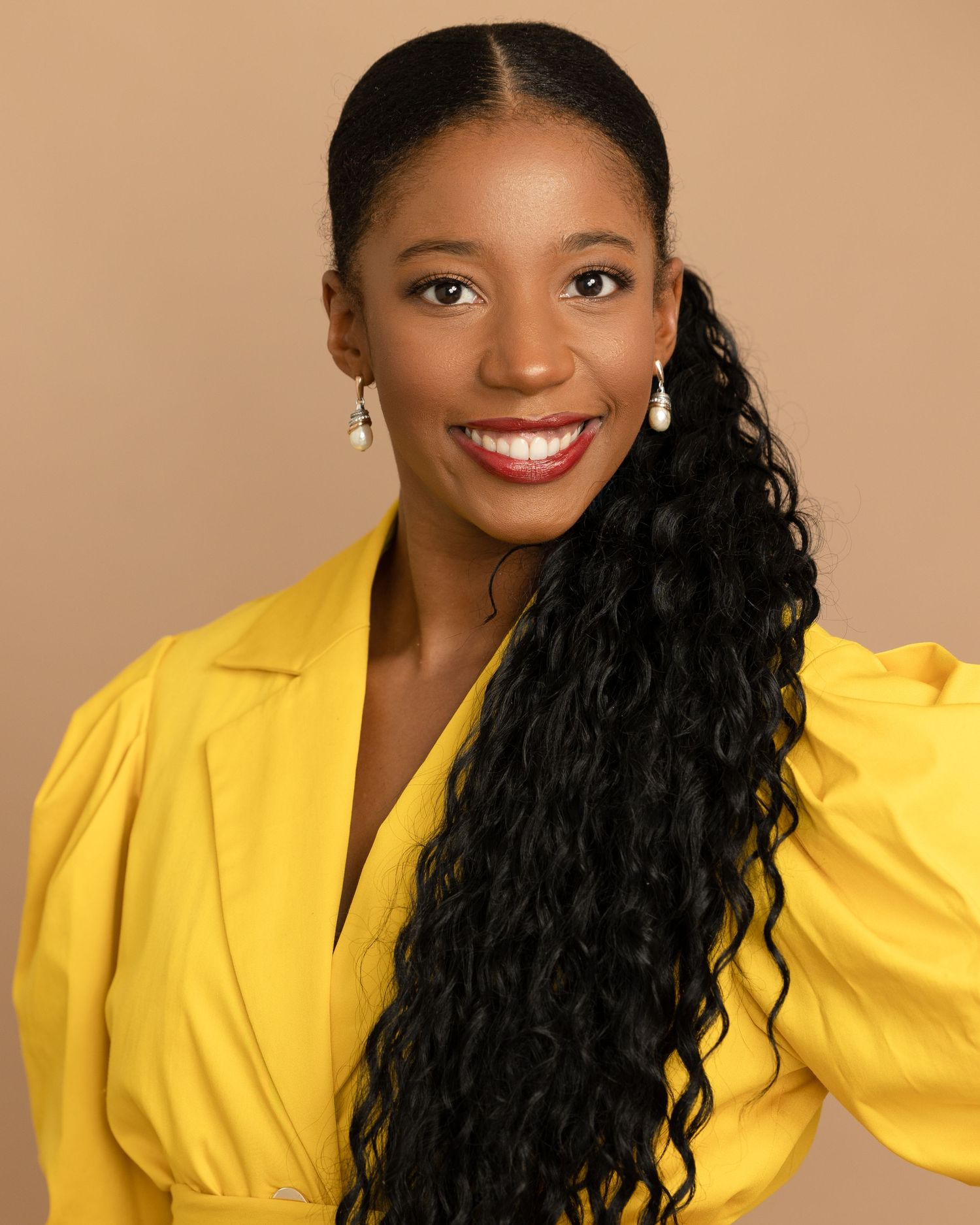 Professional portrait in bright yellow blazer against neutral background with long curly hair.