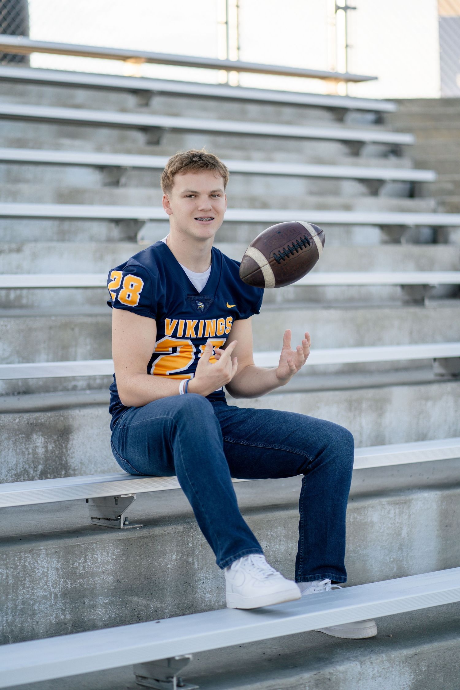 Football player in a navy Vikings jersey sits casually on bleachers tossing a football in the air.