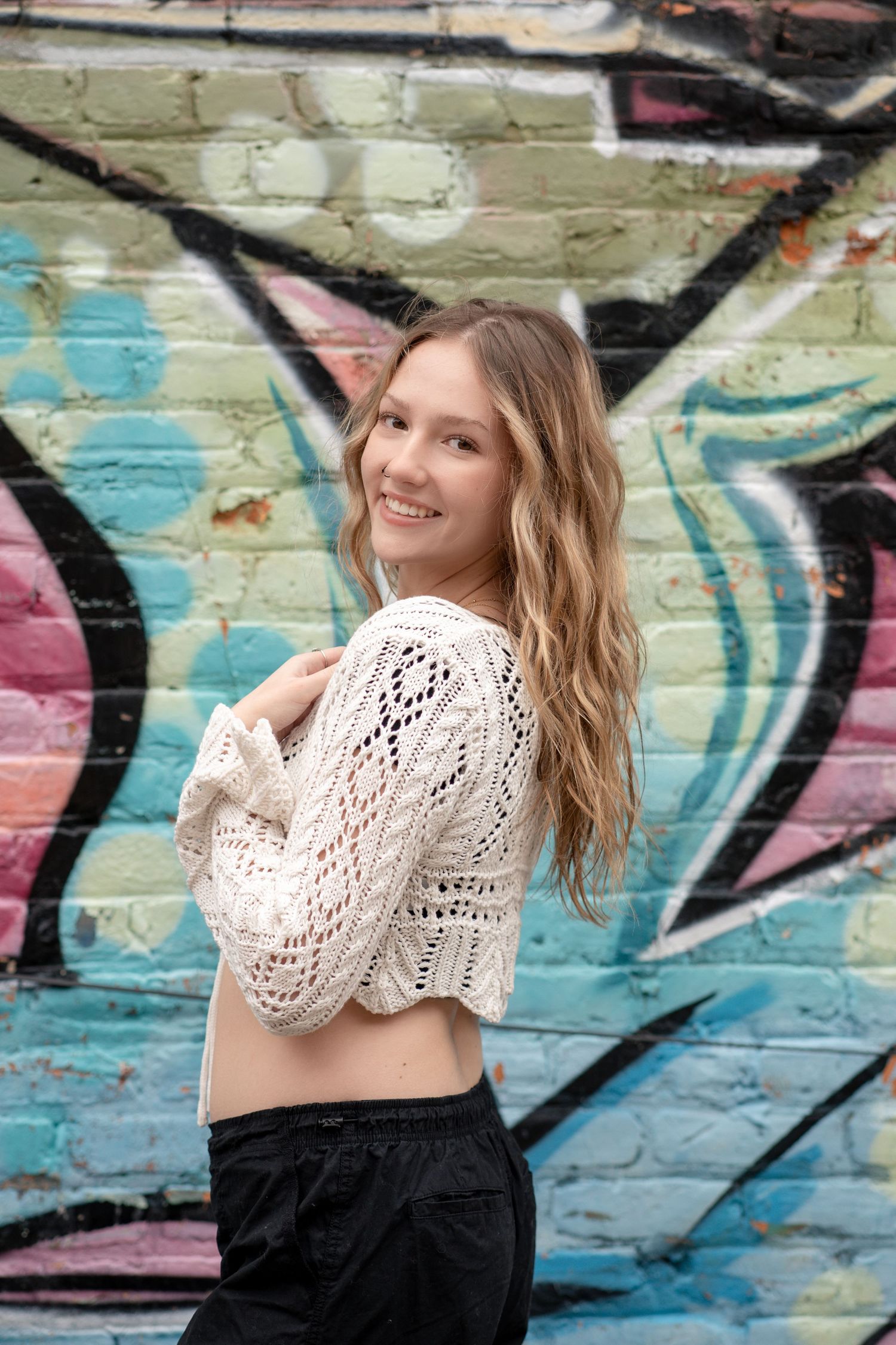 Someone in a cream knit sweater and black skirt poses playfully against a graffiti wall with swirling blue and pink designs.