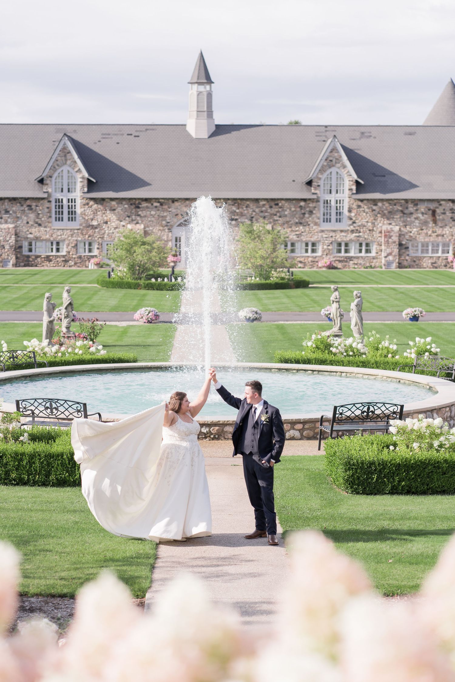 A couple celebrates their wedding in front of an elegant stone chapel with a flowing fountain and manicured gardens.