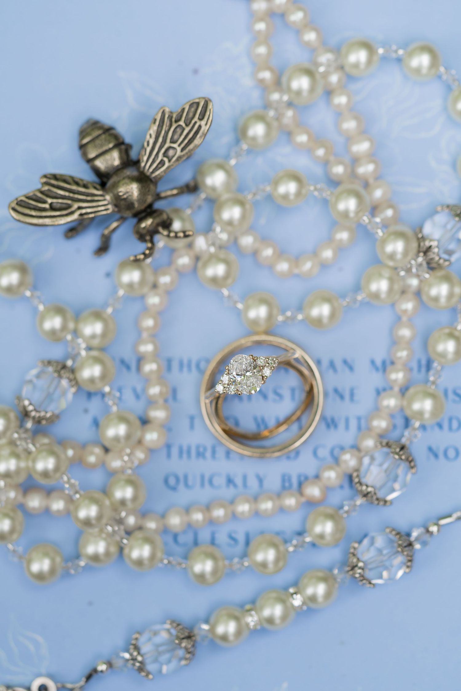 Vintage-style brass bee brooch rests among delicate pearl strands and a diamond engagement ring on blue text background.