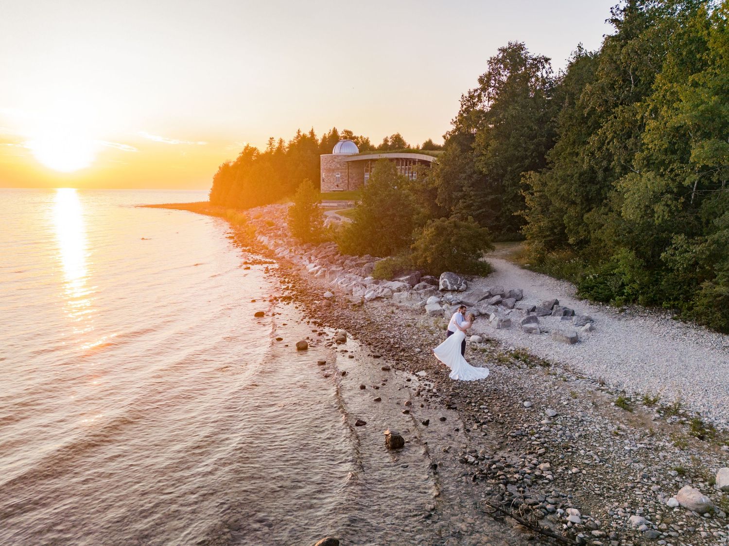Scenic sunset view of a rocky beach with a white house perched on the shore among trees near calm waters.