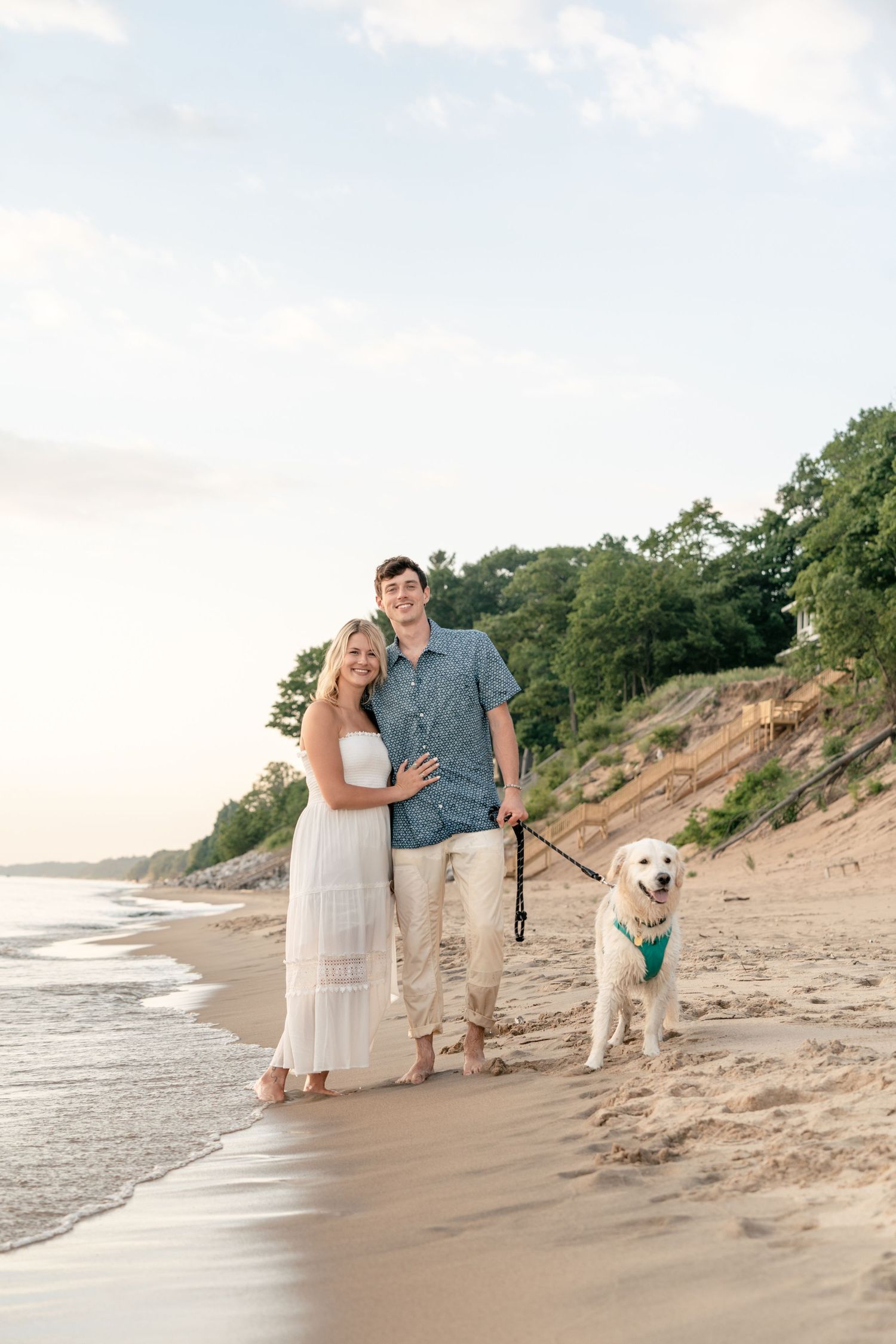 A couple and their golden retriever enjoy a romantic evening stroll along a sandy beach with eroded dunes in the background.