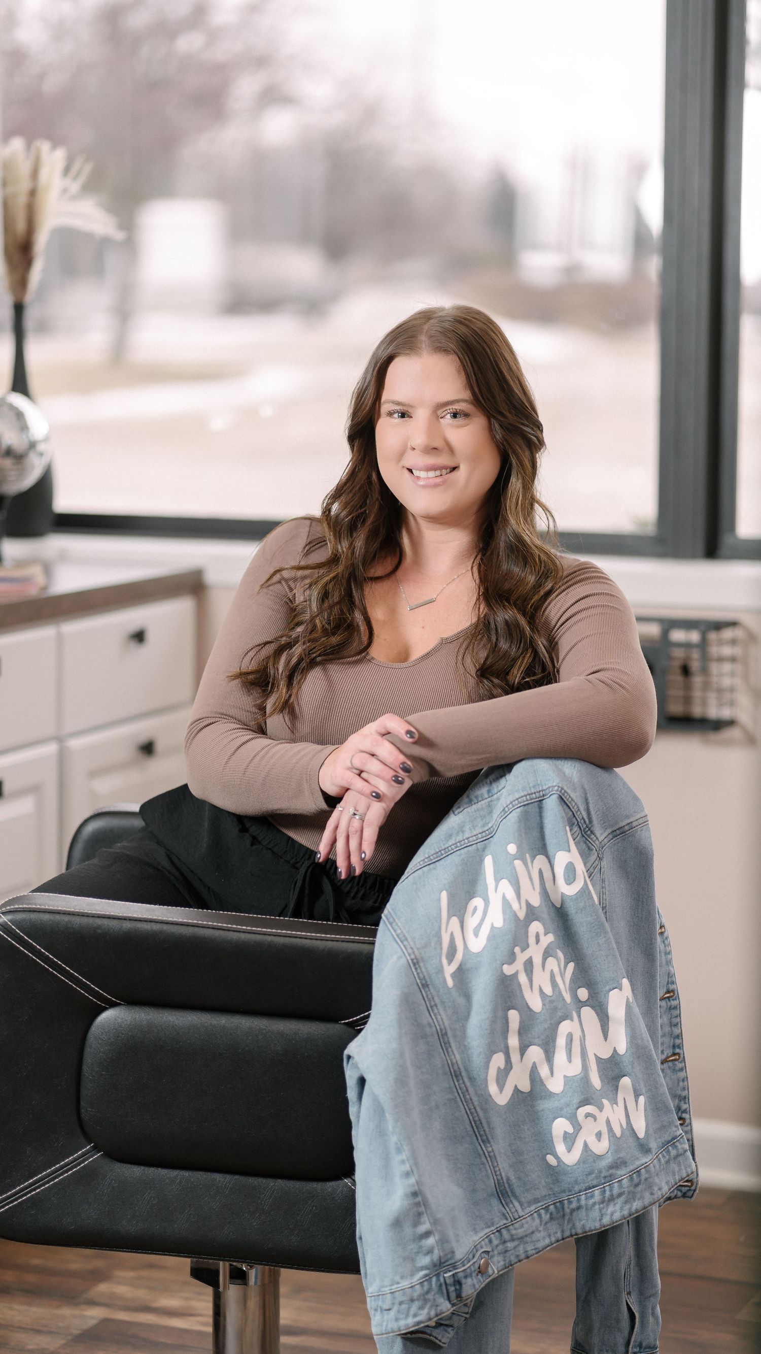 Someone sits relaxed in a salon chair holding a denim jacket with text while smiling in a modern interior space.