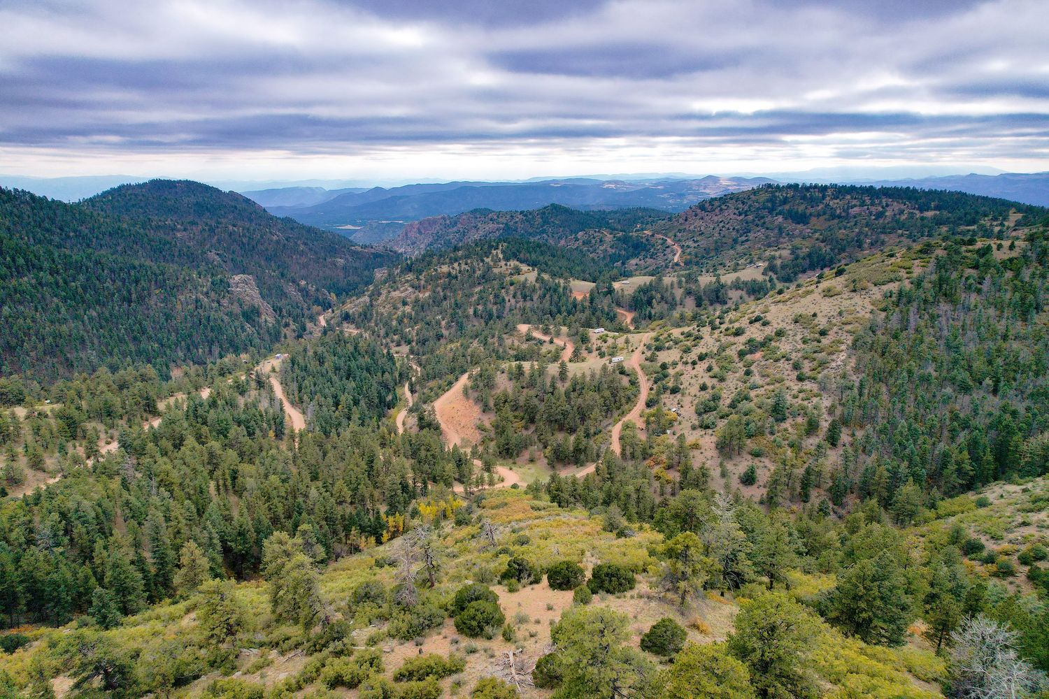 A panoramic aerial view shows winding mountain roads through forested hills and valleys under cloudy skies.