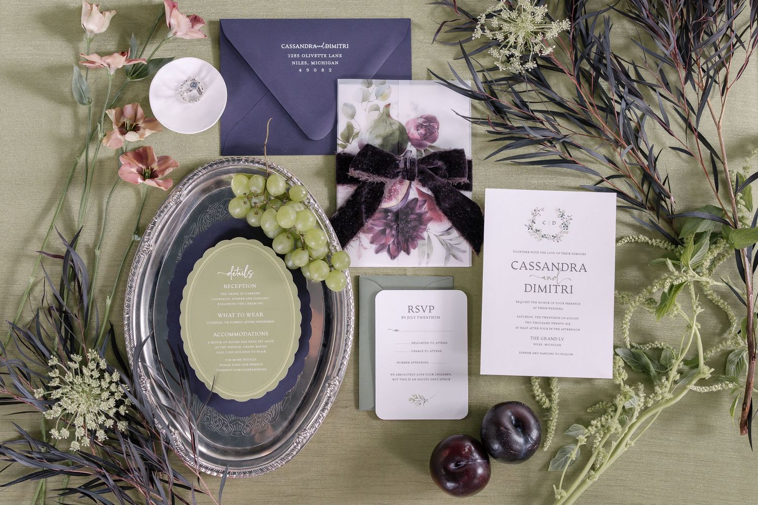 Elegant wedding invitation suite with green grapes, delicate flowers, and purple stationery on dark background.