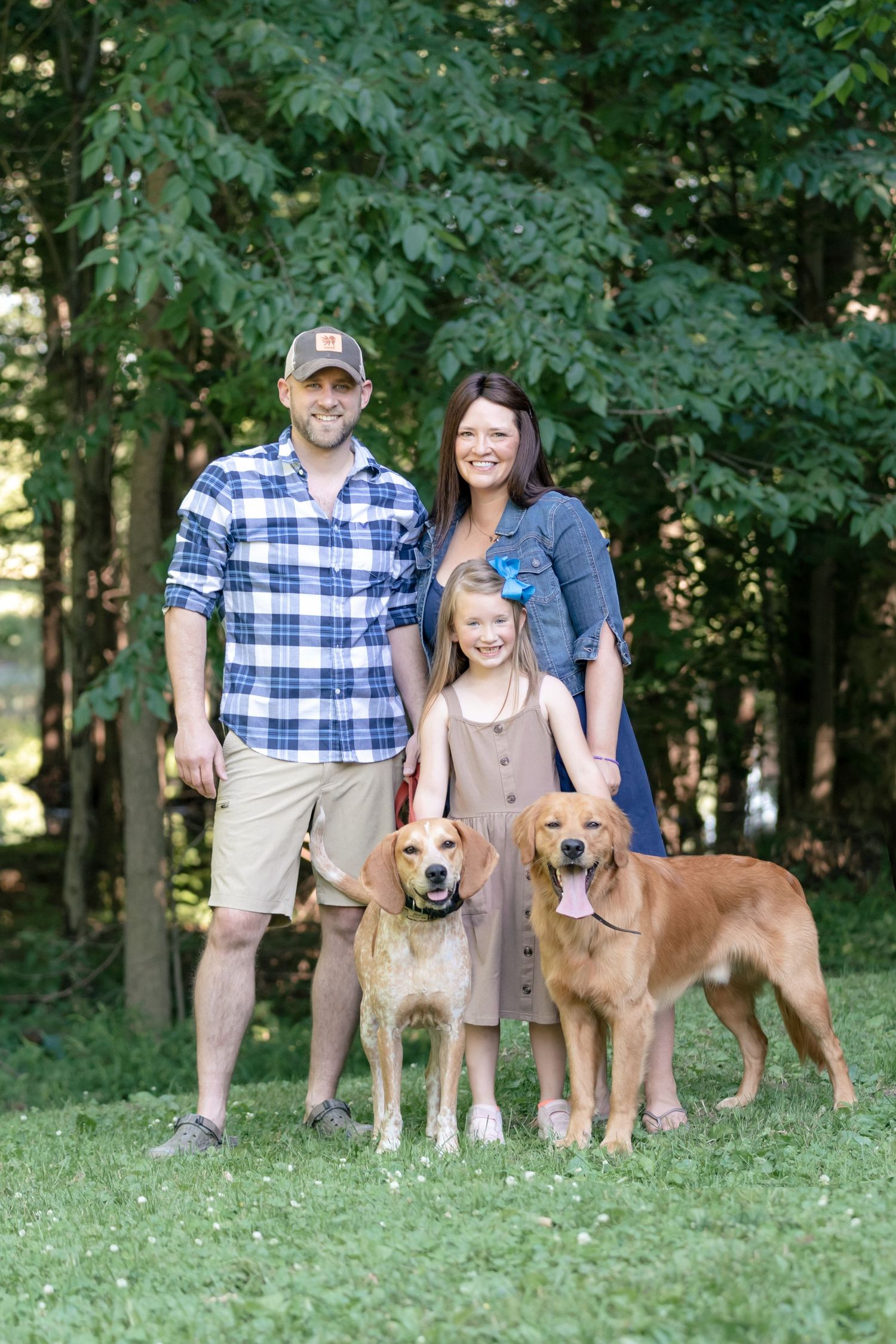 A family poses with their two golden retriever dogs on a grassy lawn with trees in the background.
