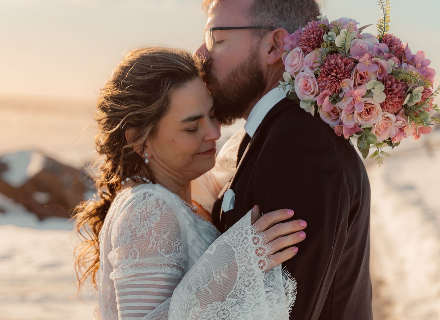 Romantic wedding couple embraces at sunset with pink floral bouquet against soft golden light.