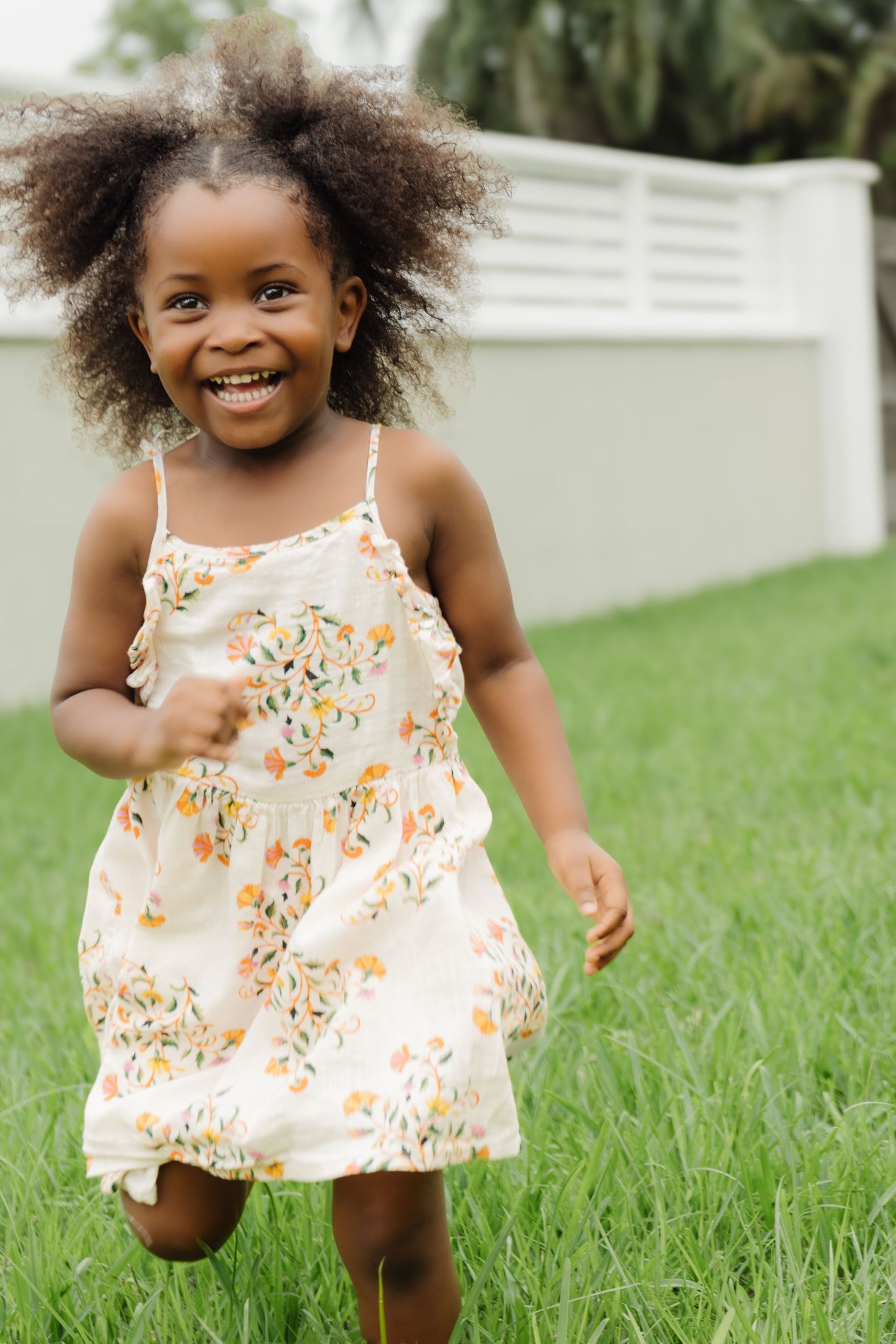 A joyful young child in a floral sundress runs and plays in a grassy backyard.