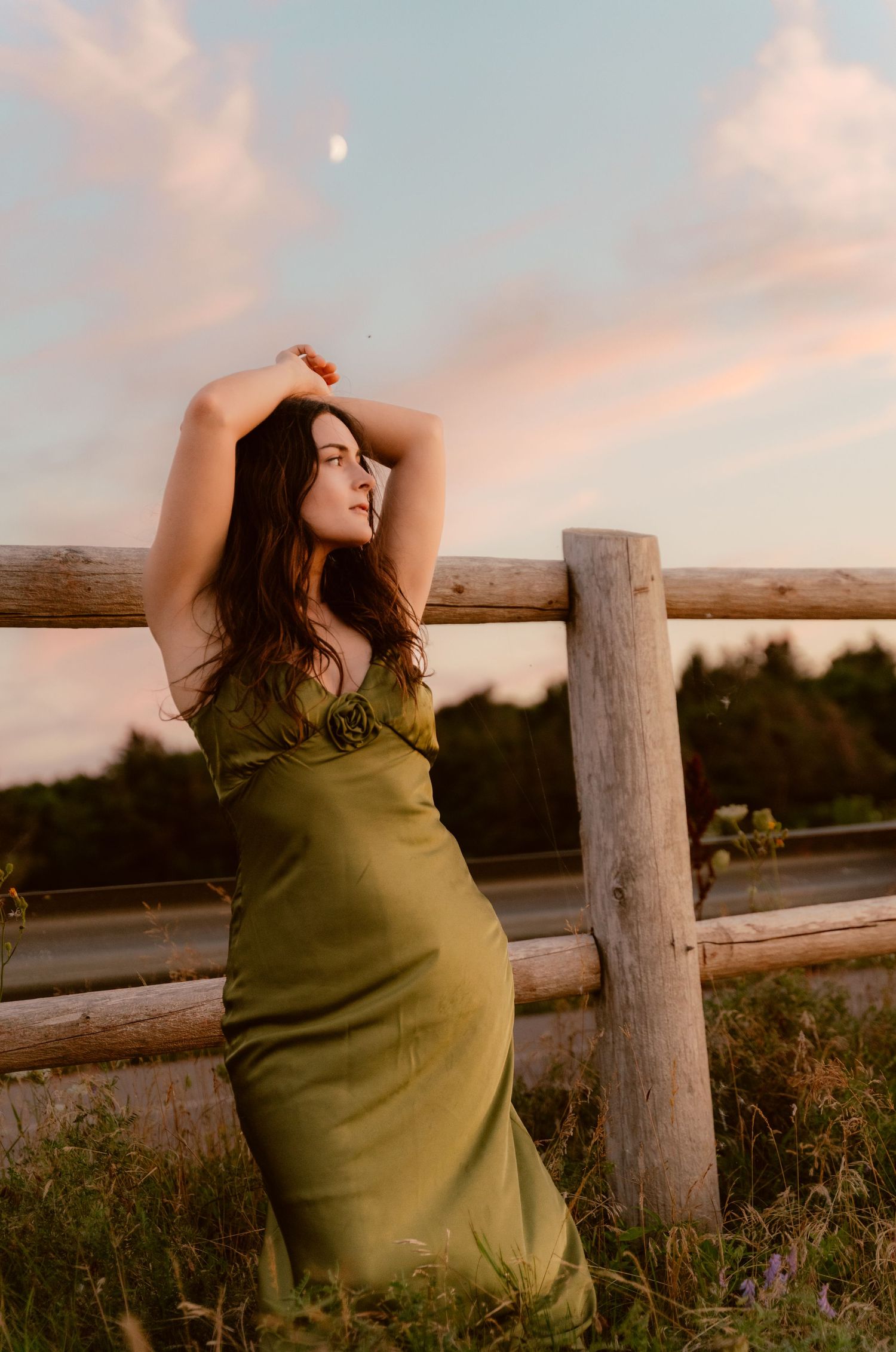 A woman in a green dress poses dramatically against a wooden fence at sunset.