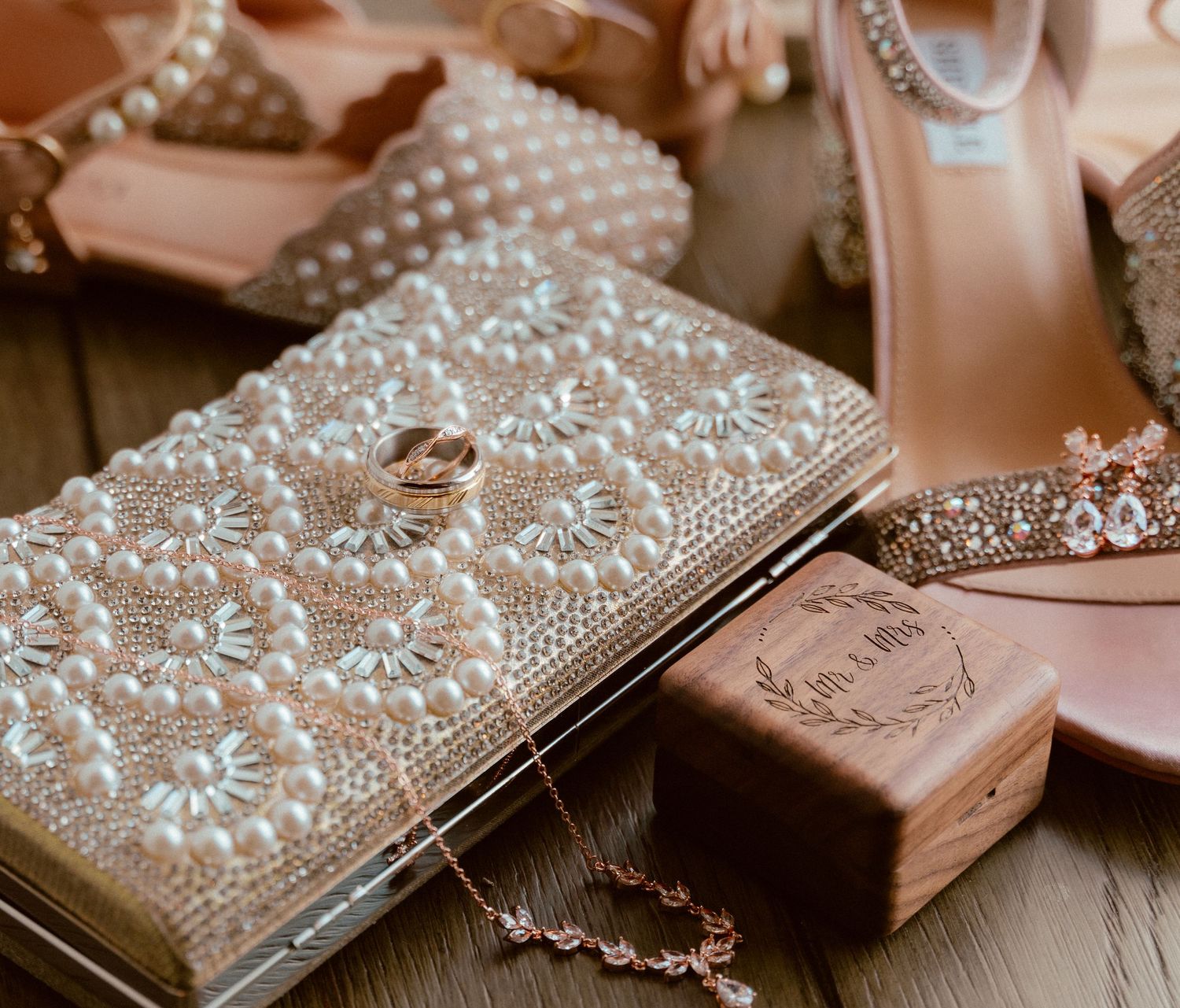 Wedding accessories including beaded clutch, rings, pink shoes and decorative items arranged on wooden surface.