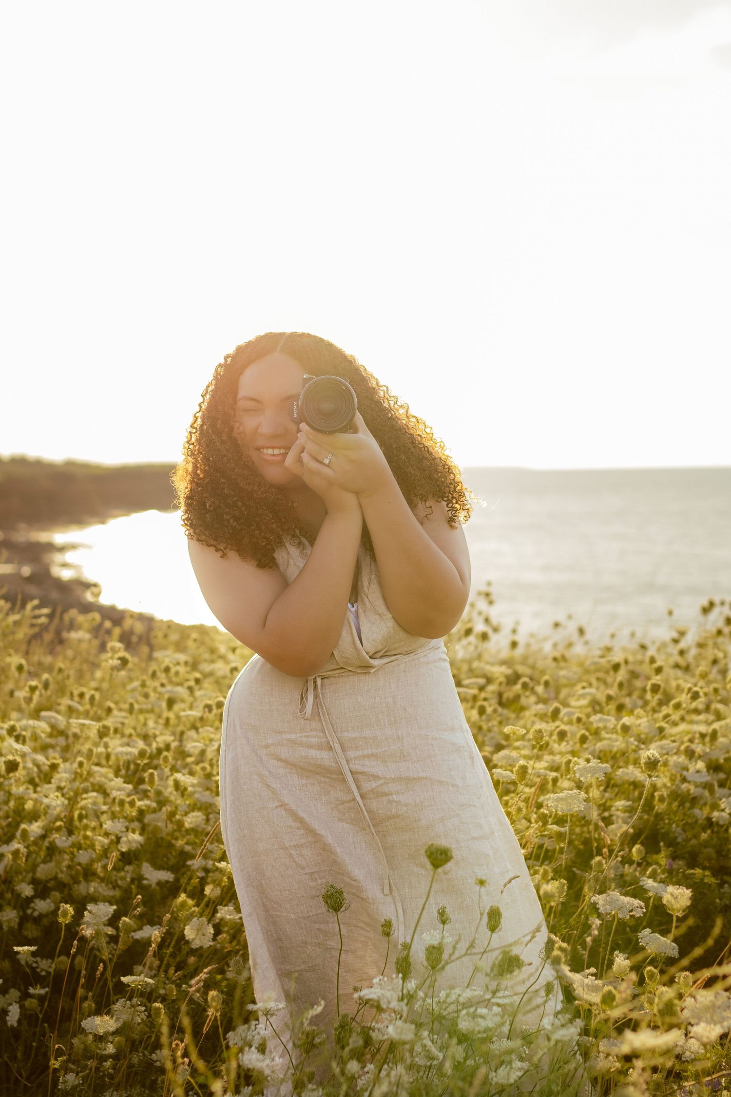 Woman in white dress laughs while standing in a golden-lit field of flowers by the sea.