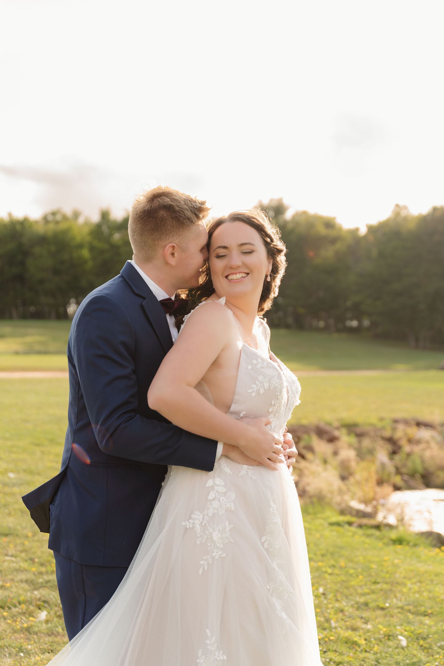 A romantic wedding couple embraces in golden sunlight on a grassy hillside during sunset.