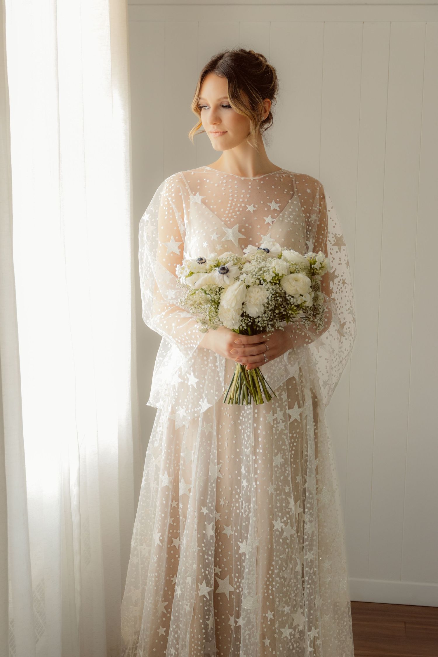 A beautiful wedding dress with sheer lace detailing and a white floral bouquet in natural light.