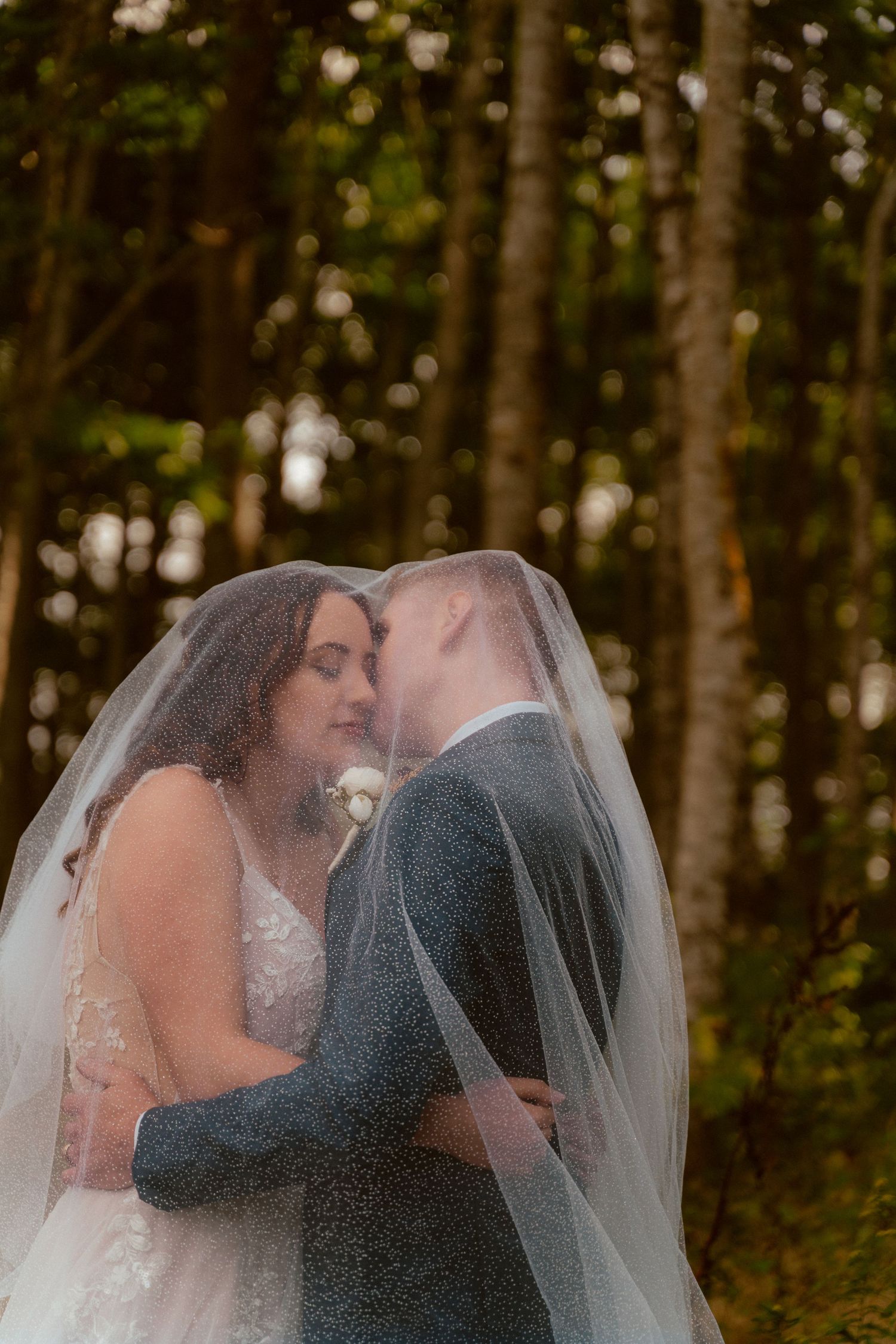 Romantic wedding couple sharing intimate moment under bridal veil in forest setting.