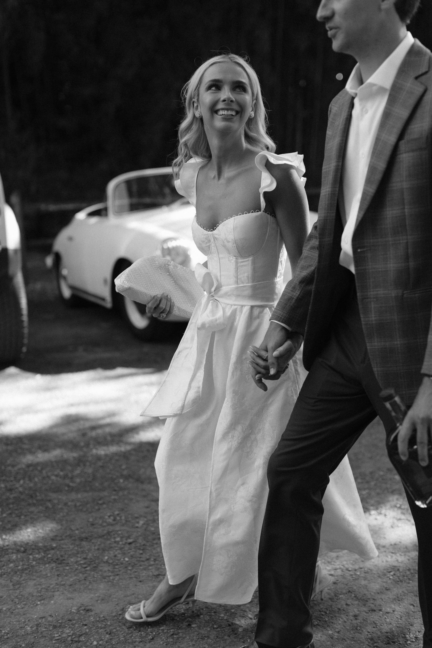 Black and white wedding photo showing a candid moment with a bride in a strapless white dress next to a classic car.