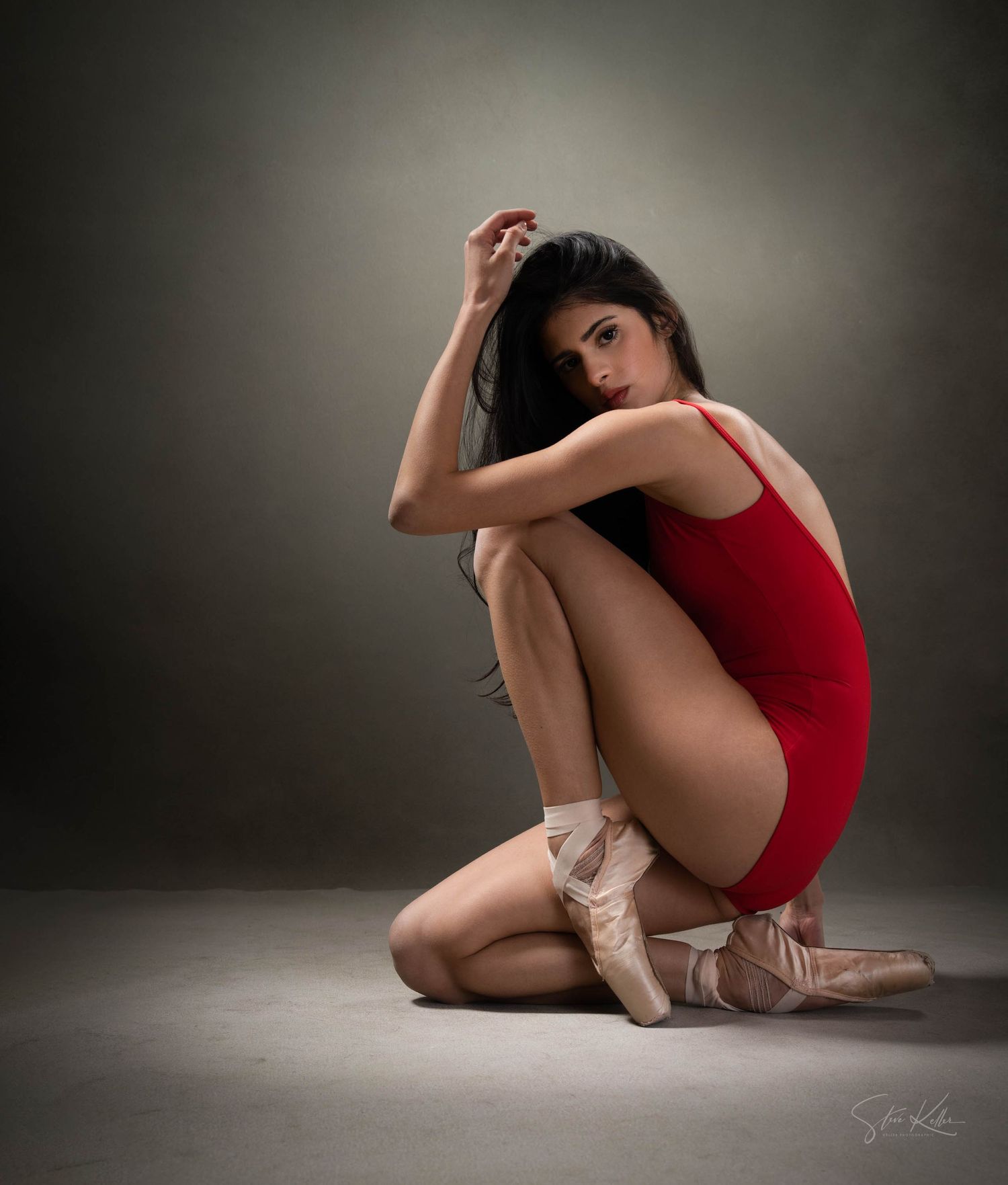Ballerina in red leotard and pointe shoes poses elegantly on studio floor.