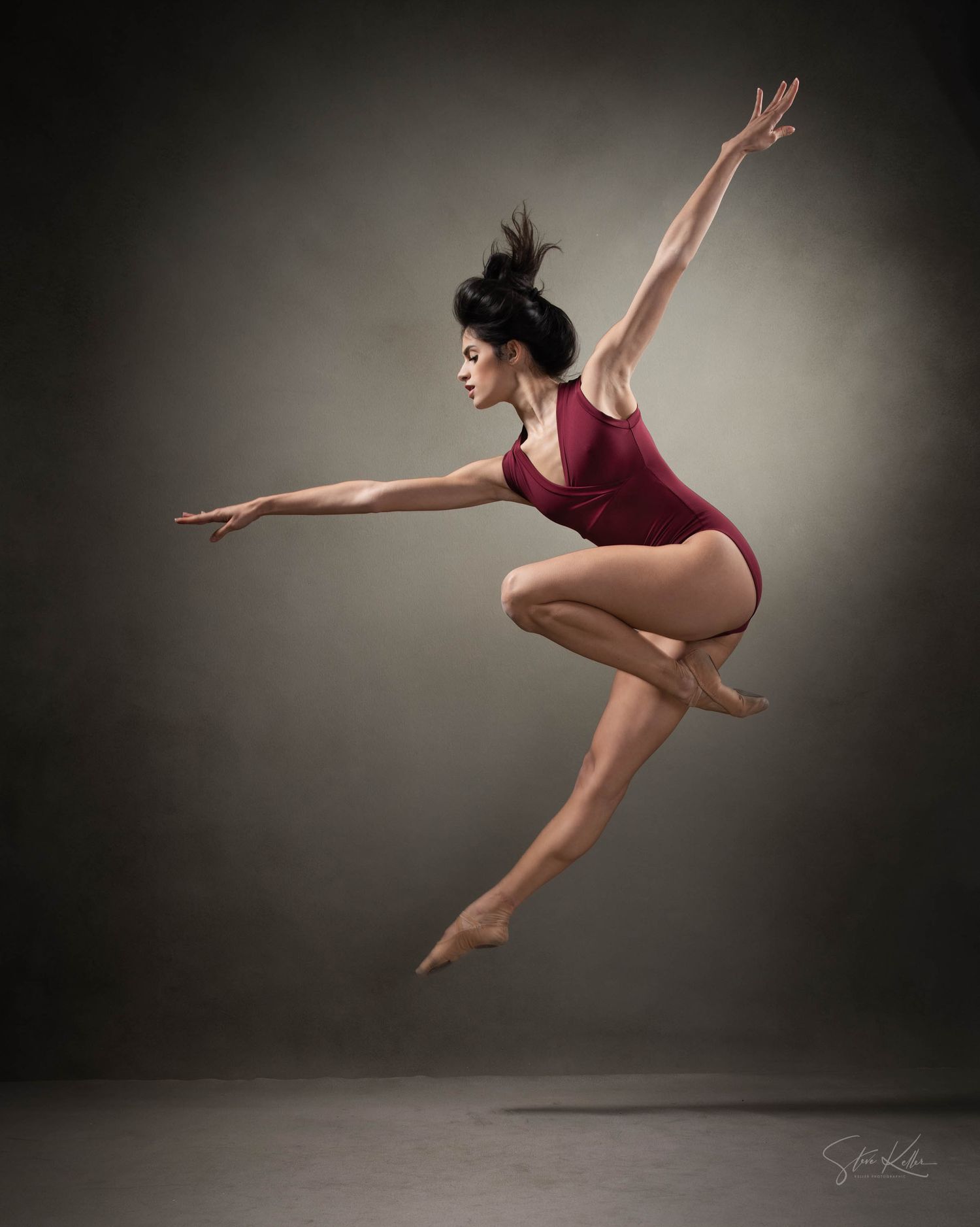 Dancer in burgundy leotard captured mid-leap with dramatic arm positioning.