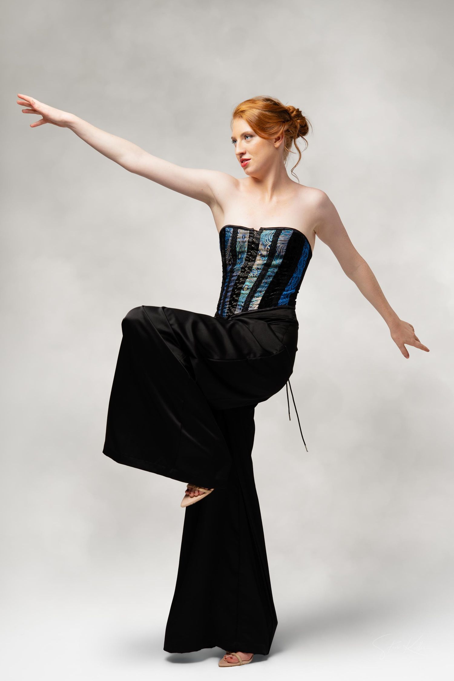 Dancer in black wide-leg pants and blue corset performs graceful pose with extended arm against gray background.