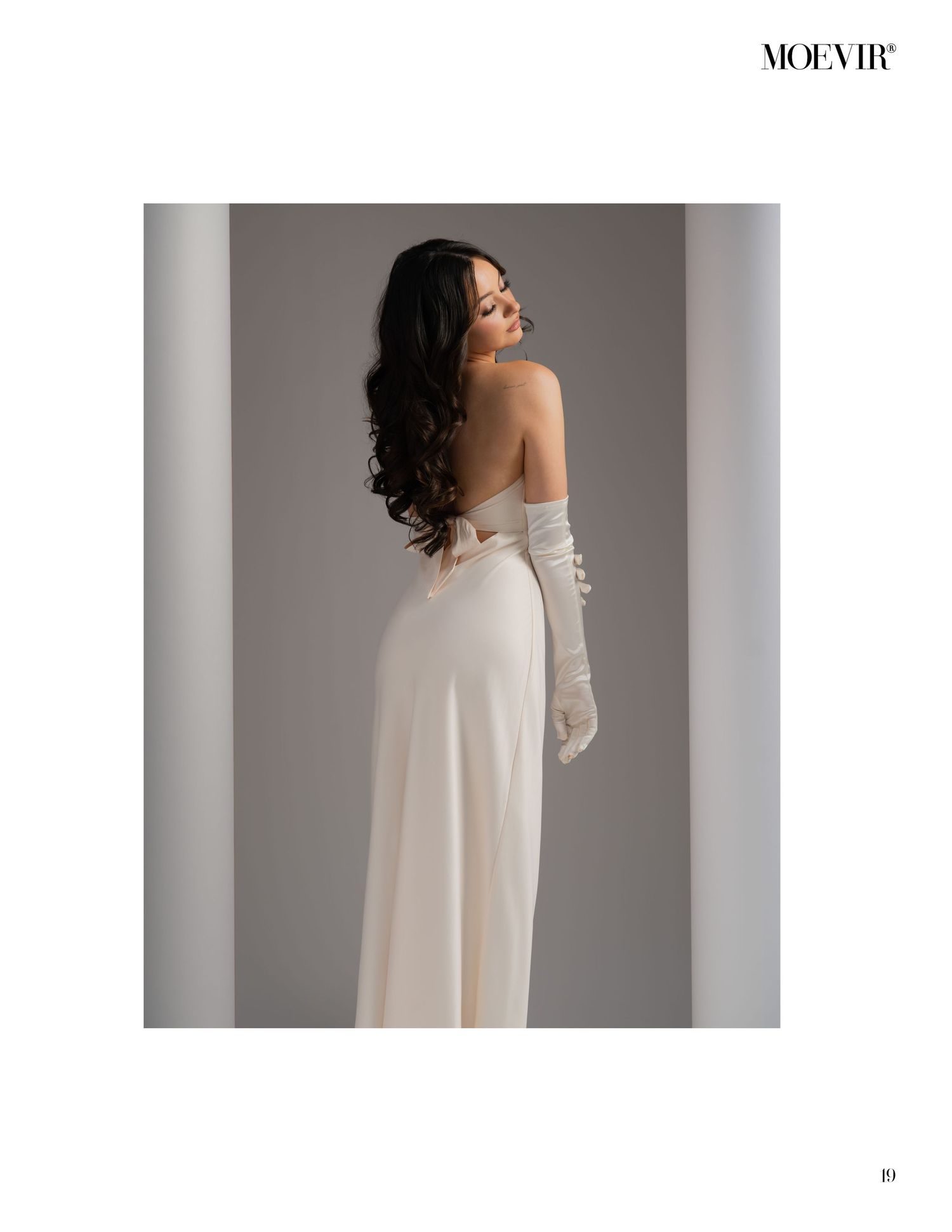 A fashion model wearing an elegant off-shoulder cream-colored gown with long gloves poses gracefully against a neutral backdrop.
