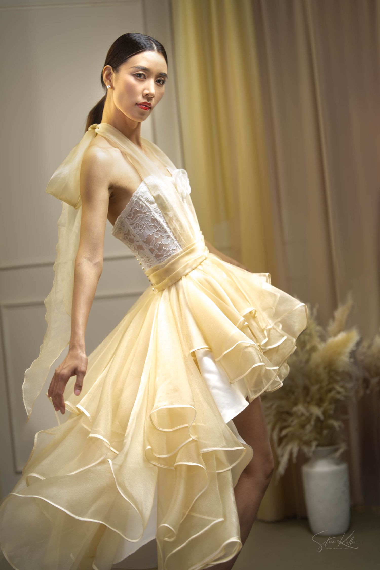 A fashion model wearing an elegant cream-colored ruffled dress with a layered skirt and fitted bodice poses in a studio setting.