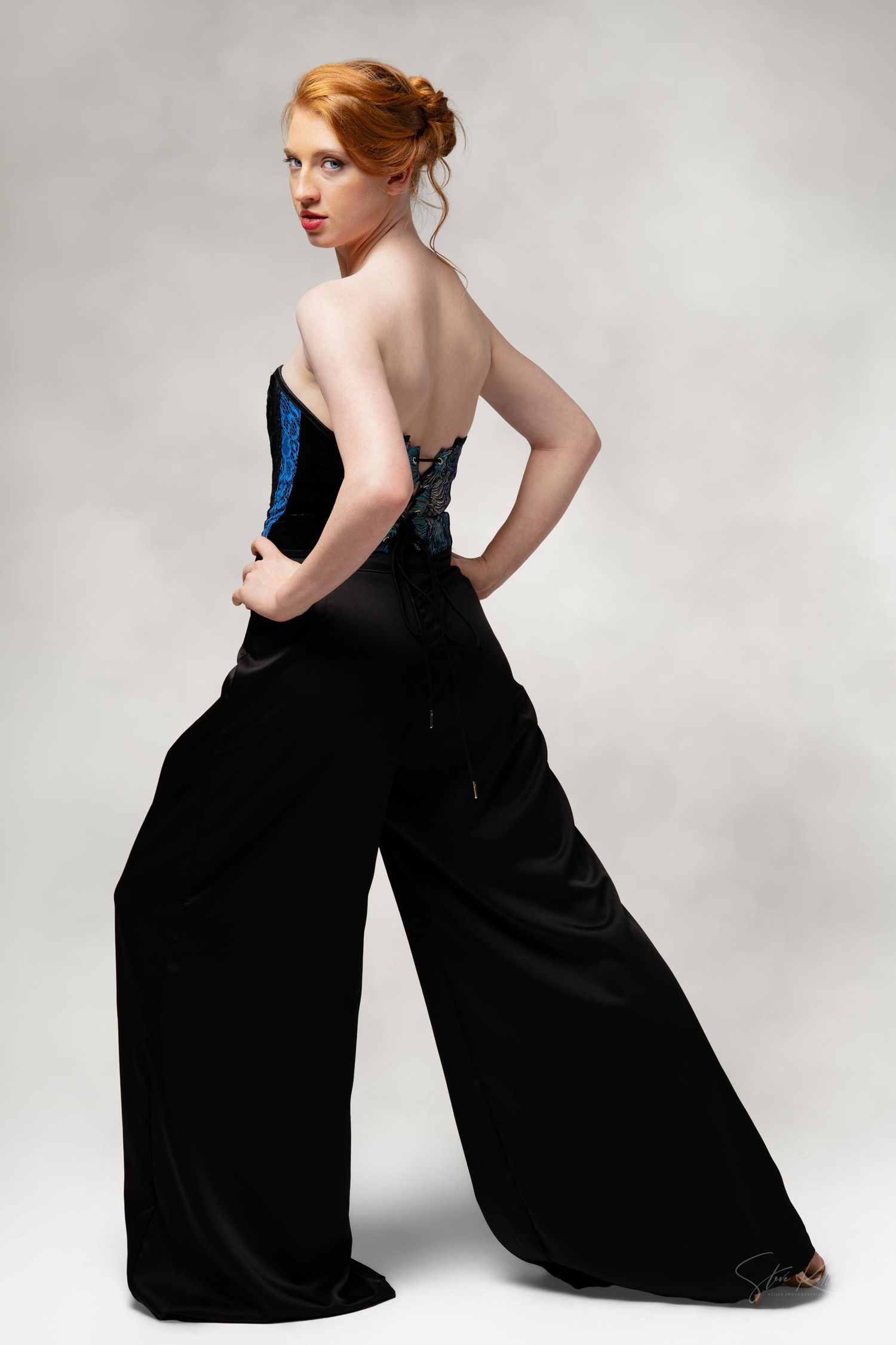 Woman in a black formal jumpsuit and blue corset poses against gray background in dramatic fashion portrait.