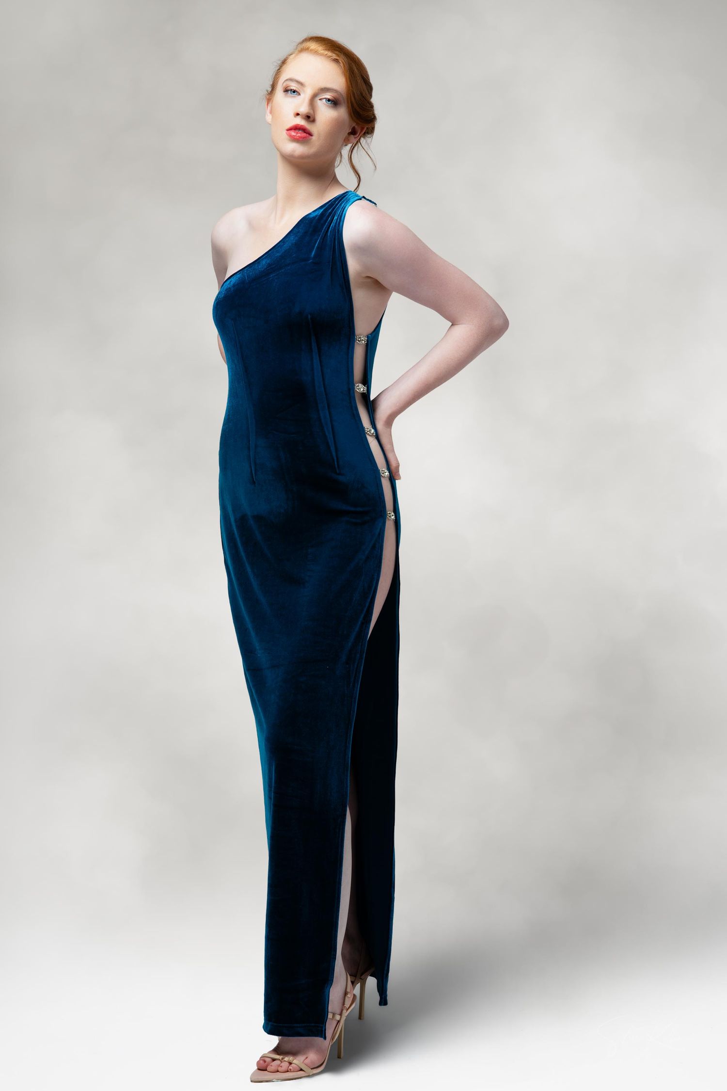 A one-shoulder navy blue velvet evening gown with high slit and metallic heels against a grey backdrop.