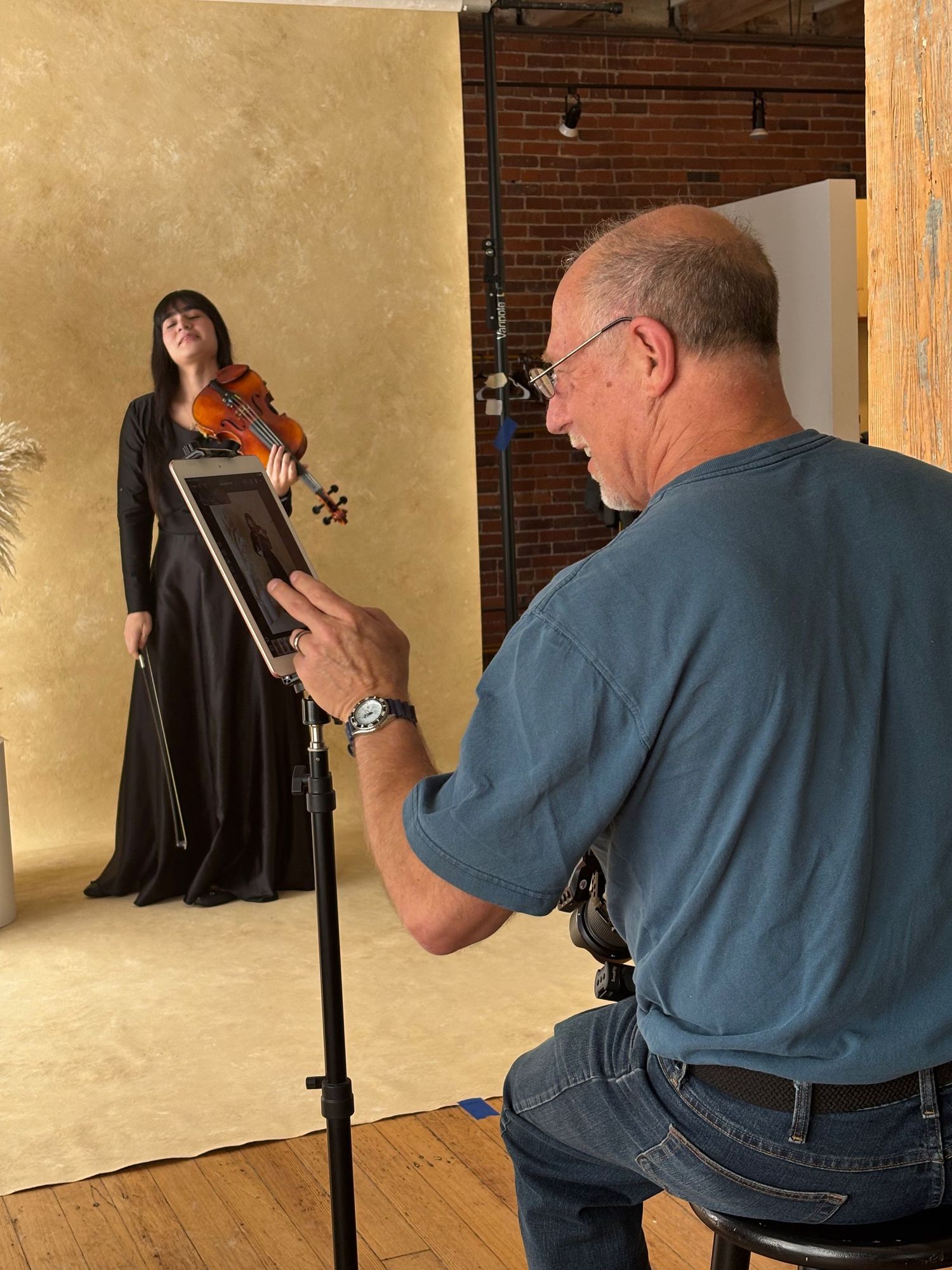 A photographer takes pictures of a violinist in a black dress against a beige backdrop in a studio setting.