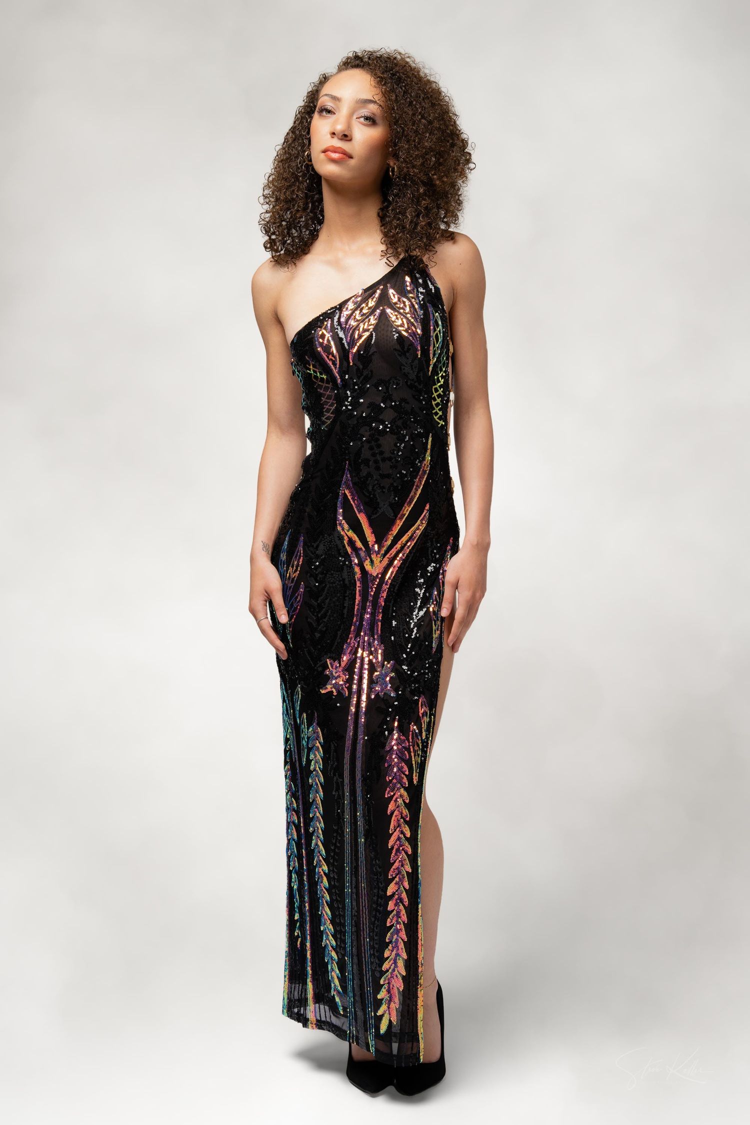 A model poses in a glamorous one-shoulder black evening gown with shimmering sequin details and geometric patterns.