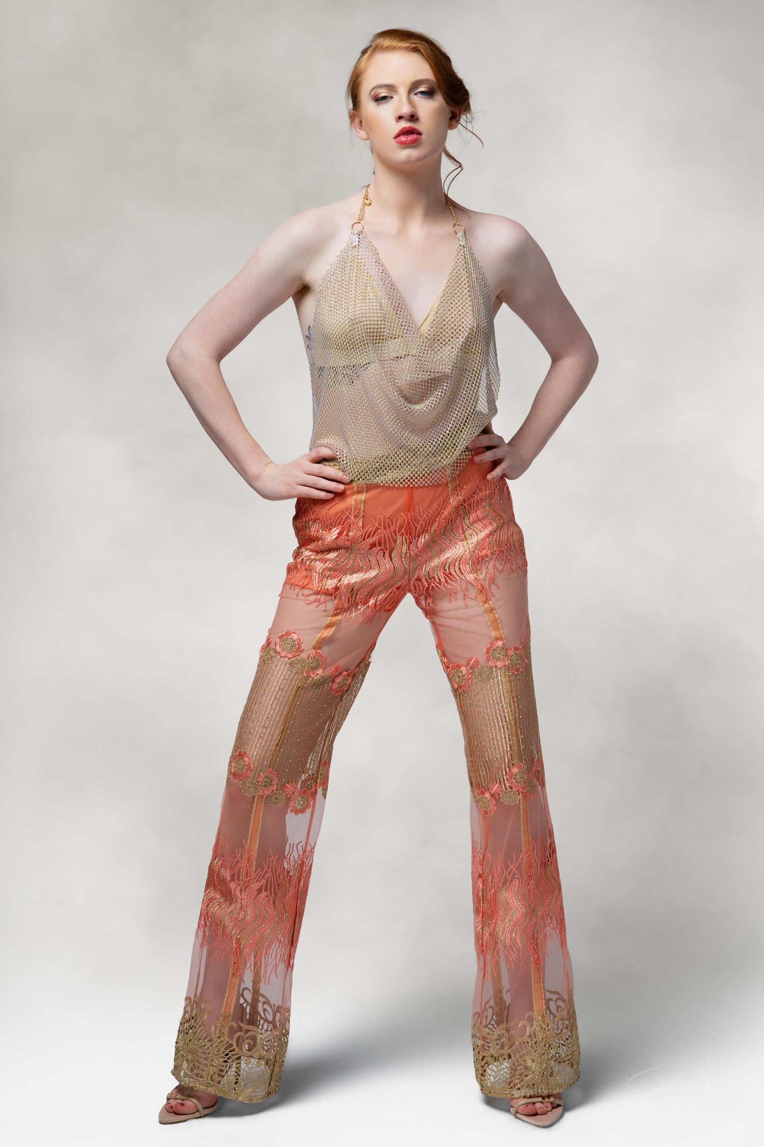 Fashion model poses in beige mesh top and coral patterned wide-leg pants against light gray background.