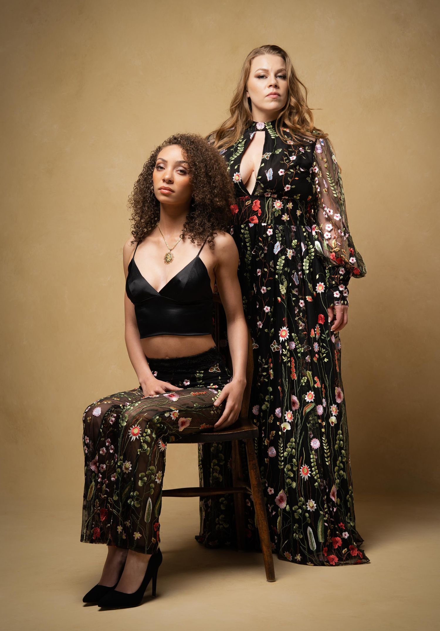 Two fashion models pose together in floral print outfits against a beige backdrop during an editorial photoshoot.