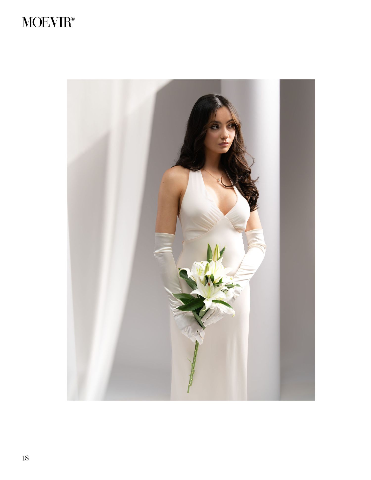 Elegant fashion model in white wedding dress and long gloves holds a single white lily stem against a minimalist light background.