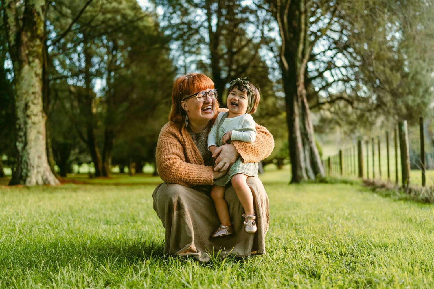 Mother and daughter laughing together outdoors, sitting on grass in a sunlit park surrounded by trees and nature.