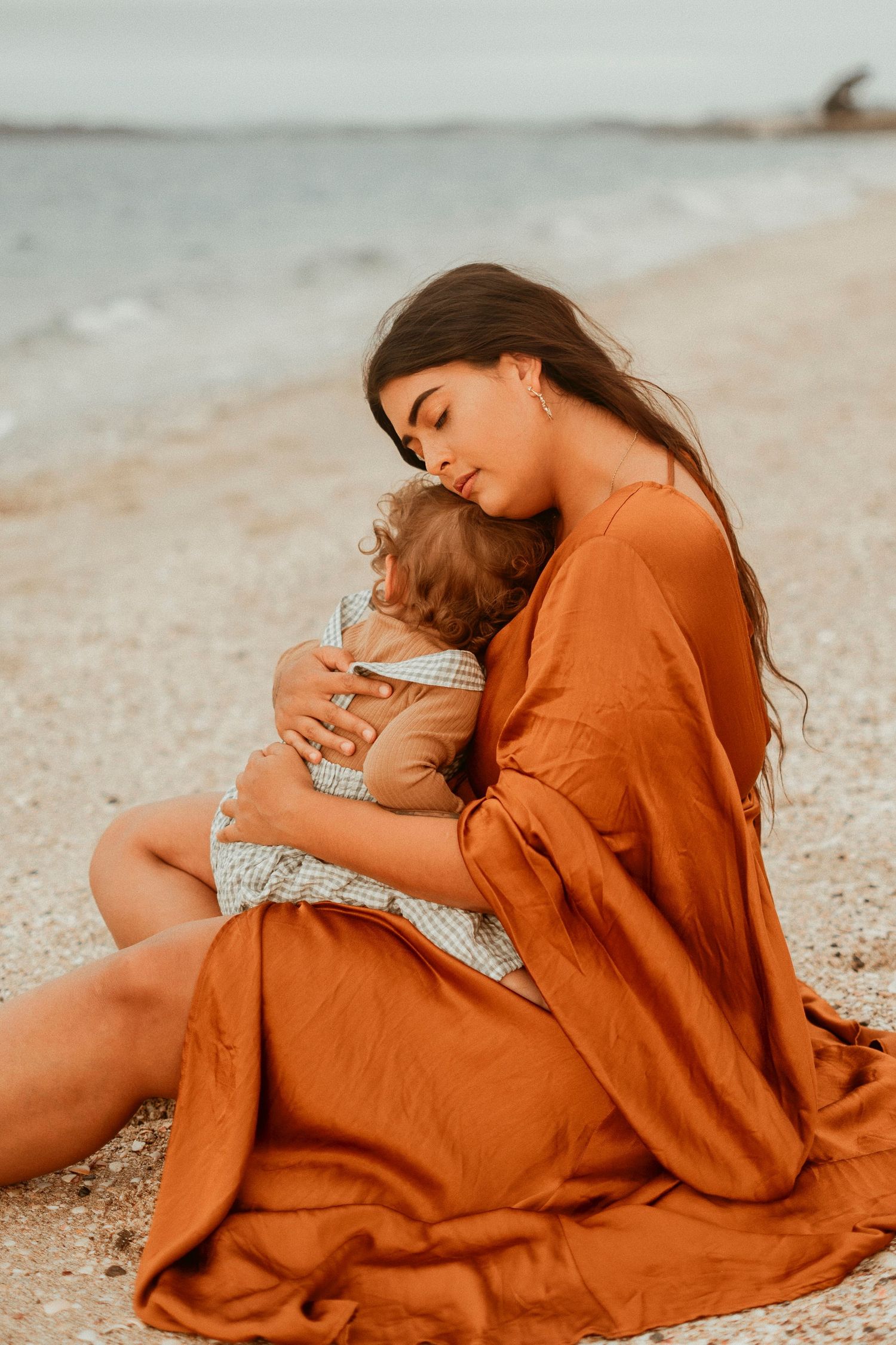 Mother in rust-colored dress cradling child on a beach, capturing a serene and intimate coastal moment.