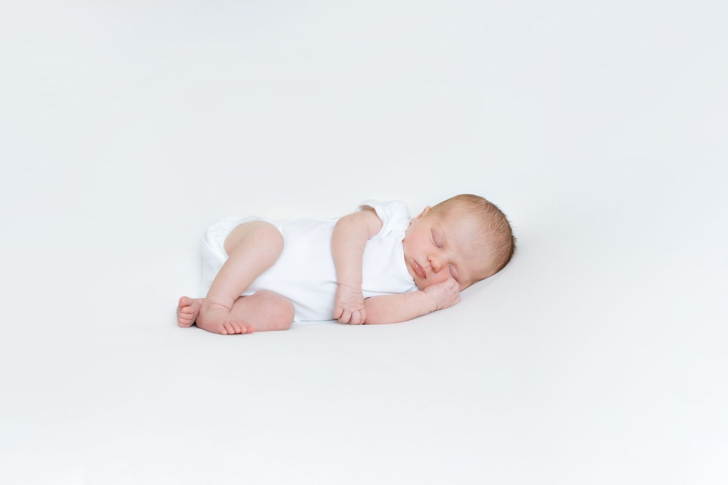 Peaceful sleeping newborn wearing white clothes curled up on a white background.
