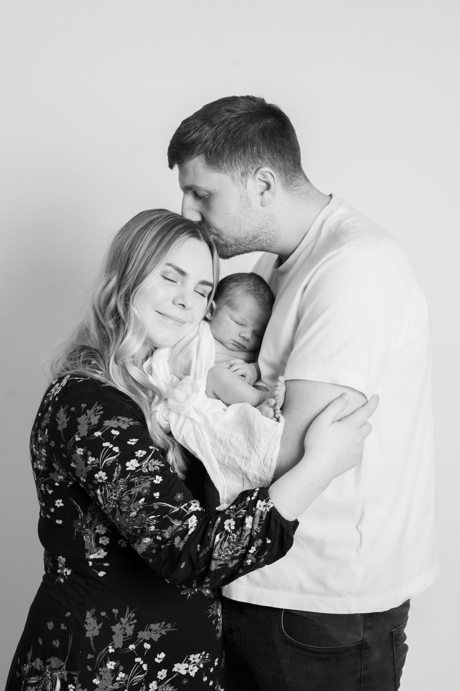 Black and white intimate portrait of parents embracing newborn baby in tender family moment.