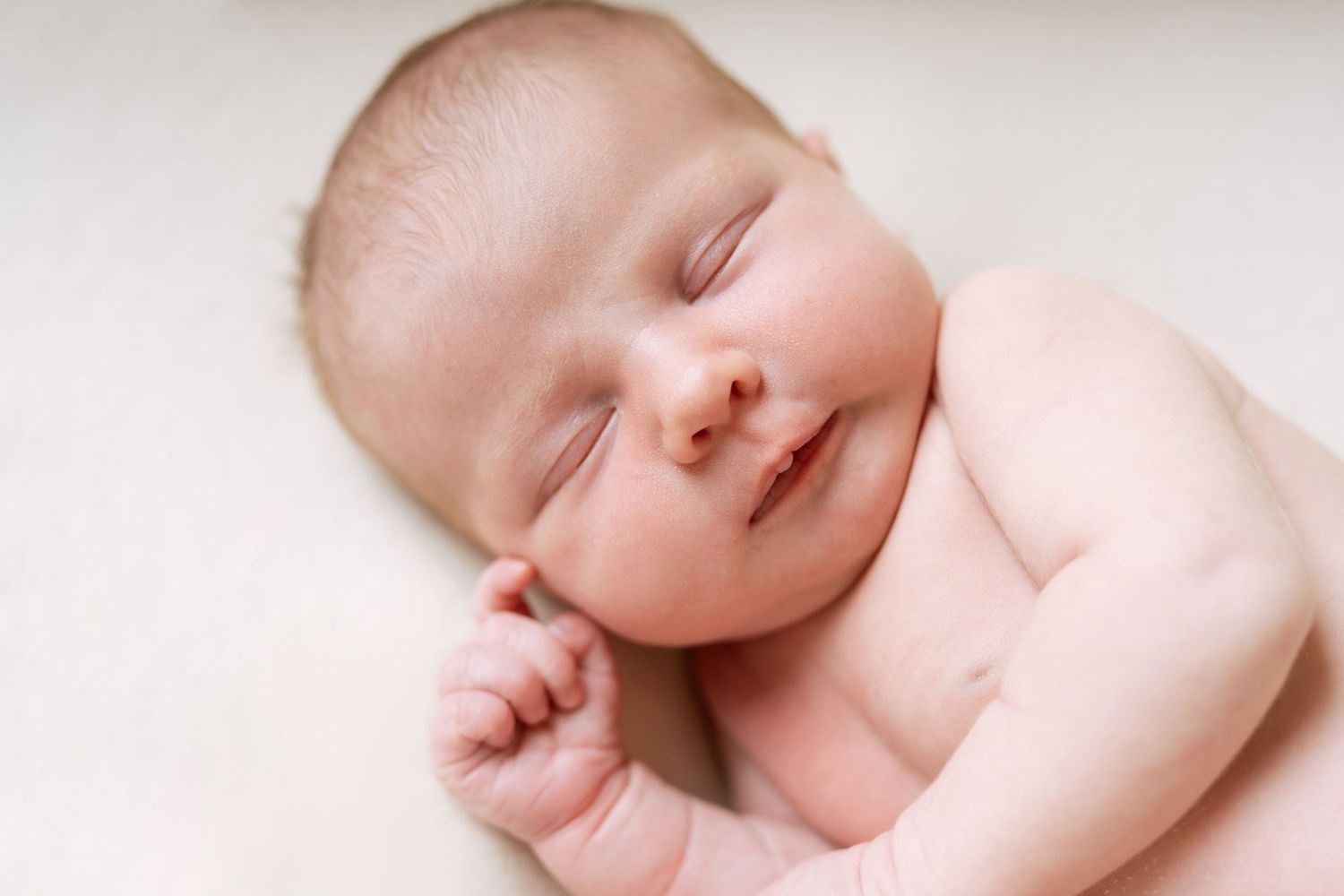 A peacefully sleeping newborn baby rests on soft beige fabric with a gentle smile and closed eyes.