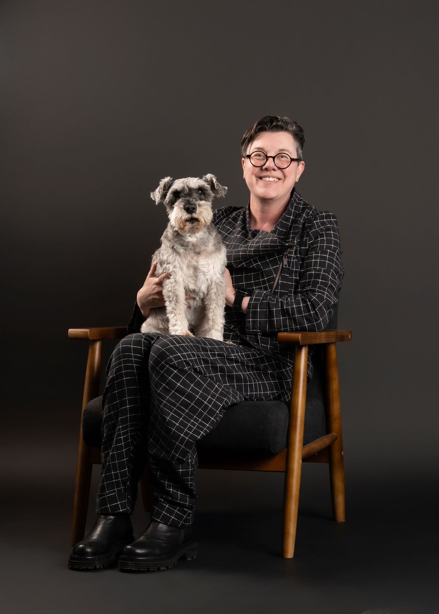 A schnauzer mix dog sits next to someone in plaid on a wooden chair against a dark background in a studio portrait.