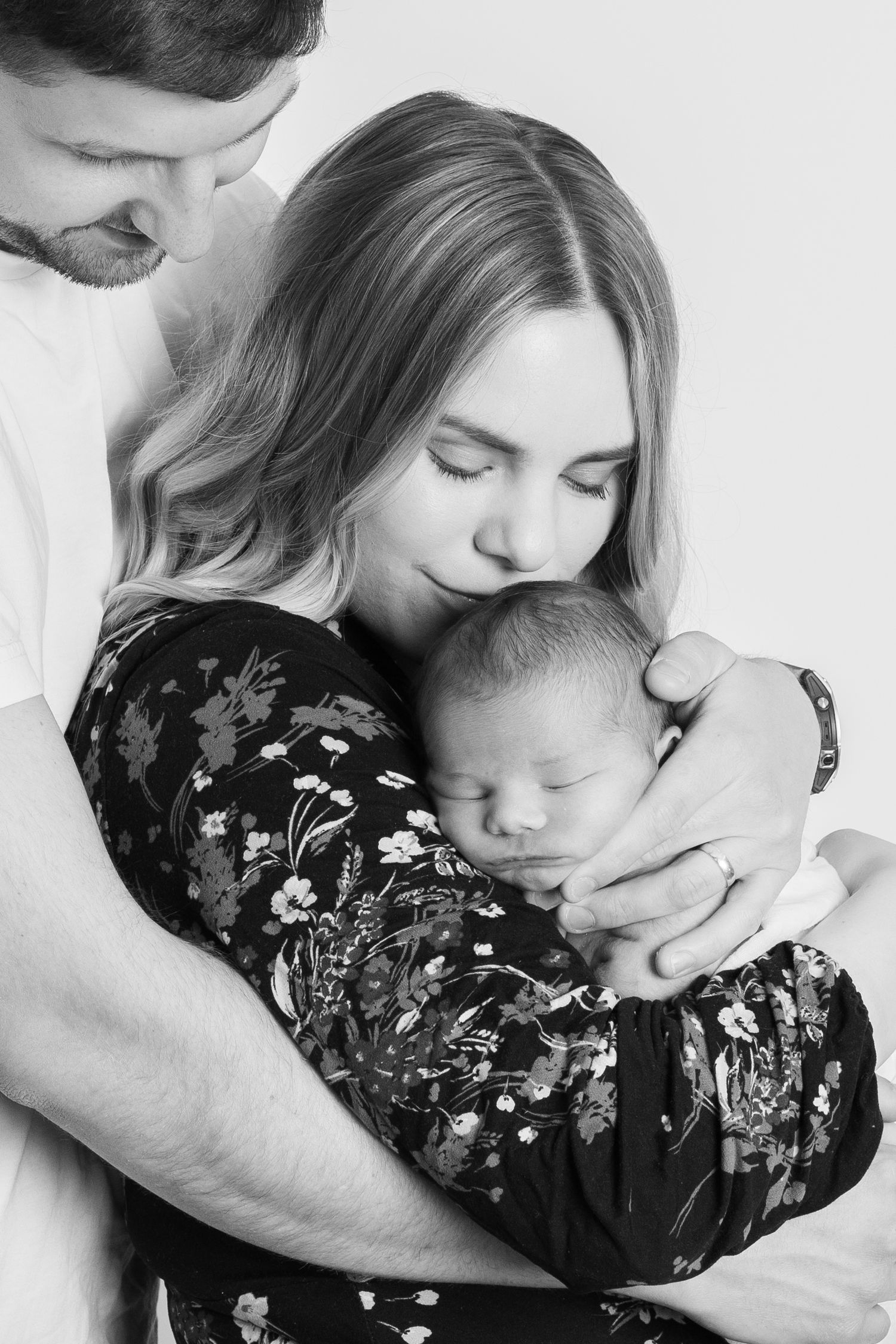 A tender black and white portrait of parents embracing their sleeping newborn baby while wearing a floral dress.